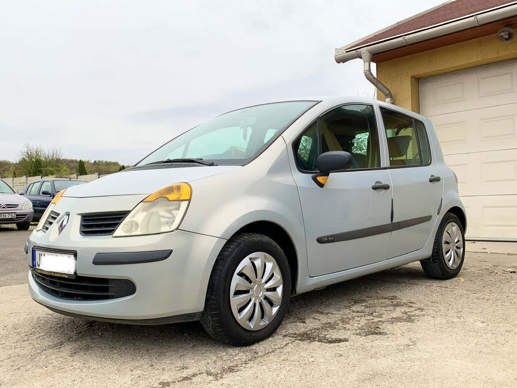Renault Modus 桌布 壁紙 — 一輛銀色雷諾Koleos停泊在碎石路上，前面是米色房屋和白色車庫門，天空多雲。