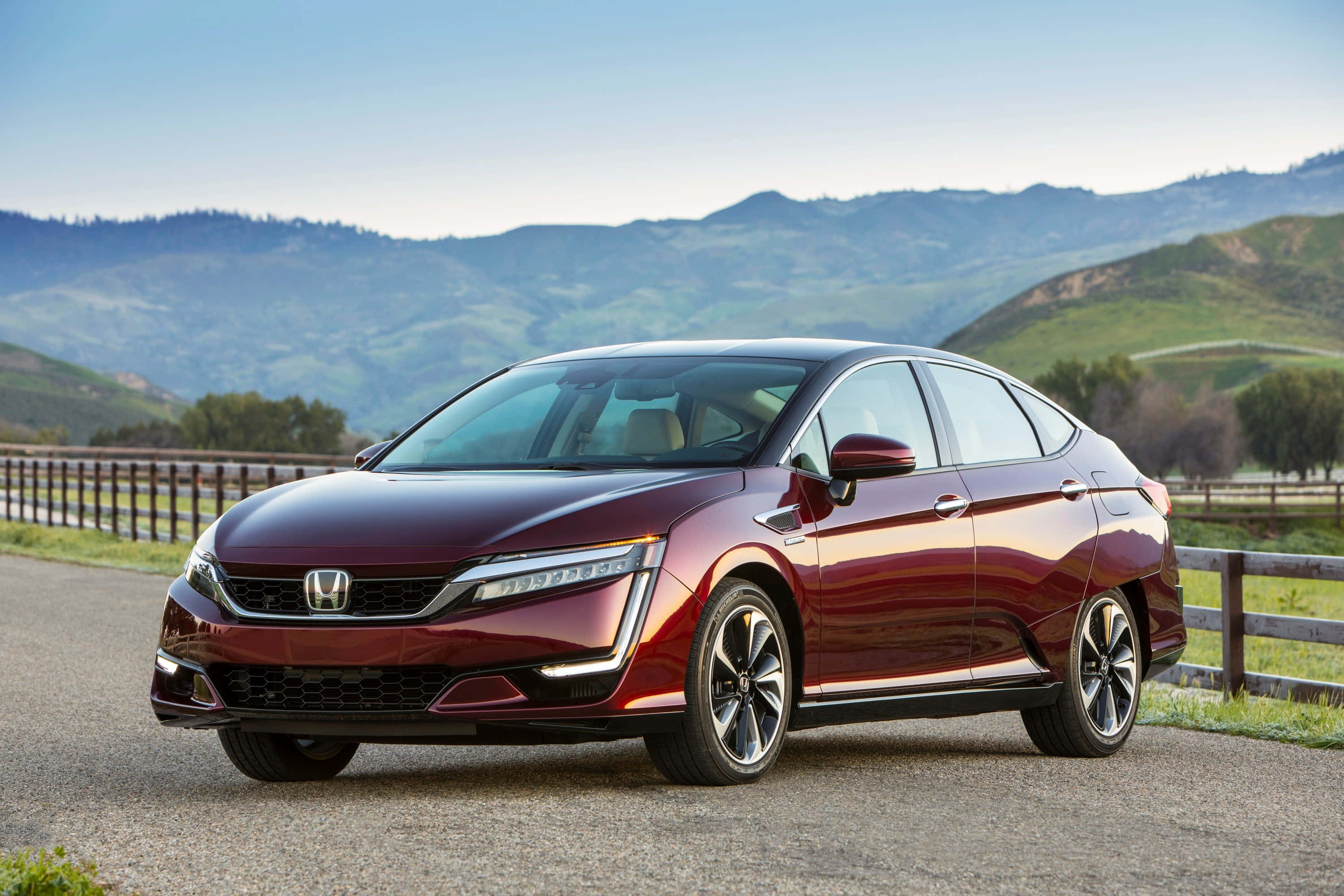 Honda Clarity 桌布 壁紙 — 一輛時尚的酒紅色本田轎車在碎石路上閃耀，背景是連綿的綠色山丘和木柵欄，天空清澈湛藍。
