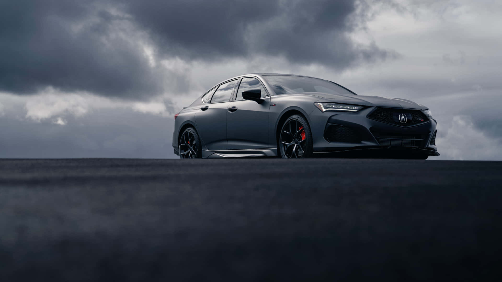 Acura Tlx 桌布 壁紙 — 一輛時尚的啞光灰色運動轎車配備紅色制動卡鉗，在陰沉暴風雨的天空下矗立在黑暗的平原上。
