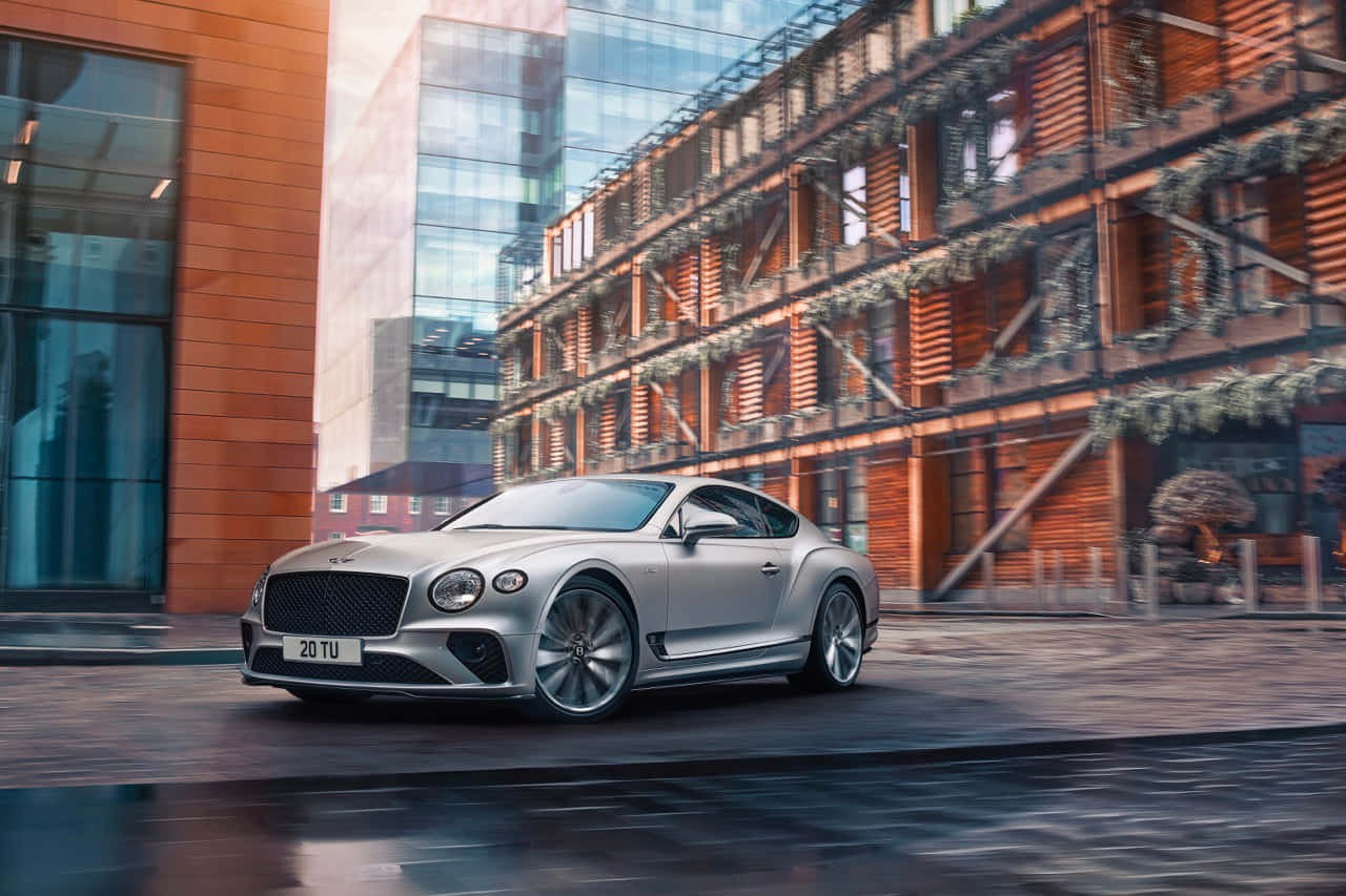 Bentley Continental Gt 桌布 壁紙 — 一輛時尚的銀色賓利Continental GT在柔和日光下行駛於現代城市街道，兩旁是玻璃和磚砌建築。