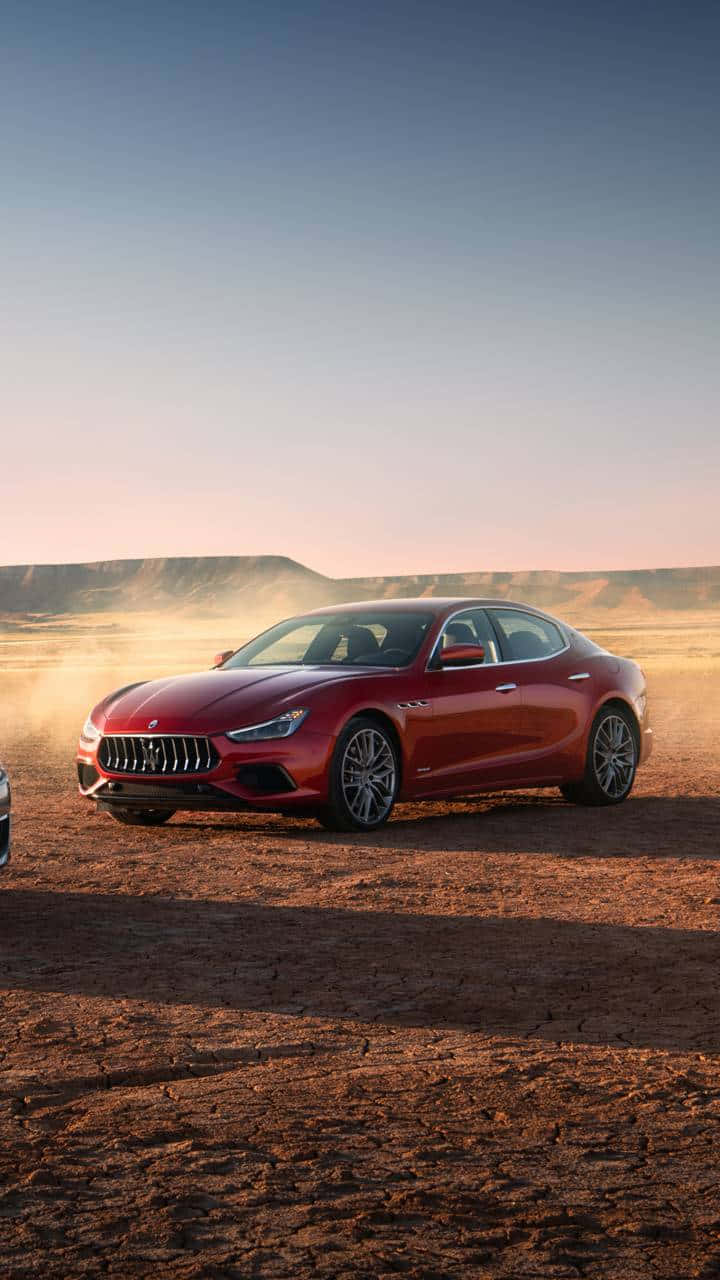 Maserati Ghibli 桌布 壁紙 — 一輛時尚的紅色Maserati轎車在黃金時段滑過塵土飛揚的沙漠景觀，溫暖的光線投下長長的影子。