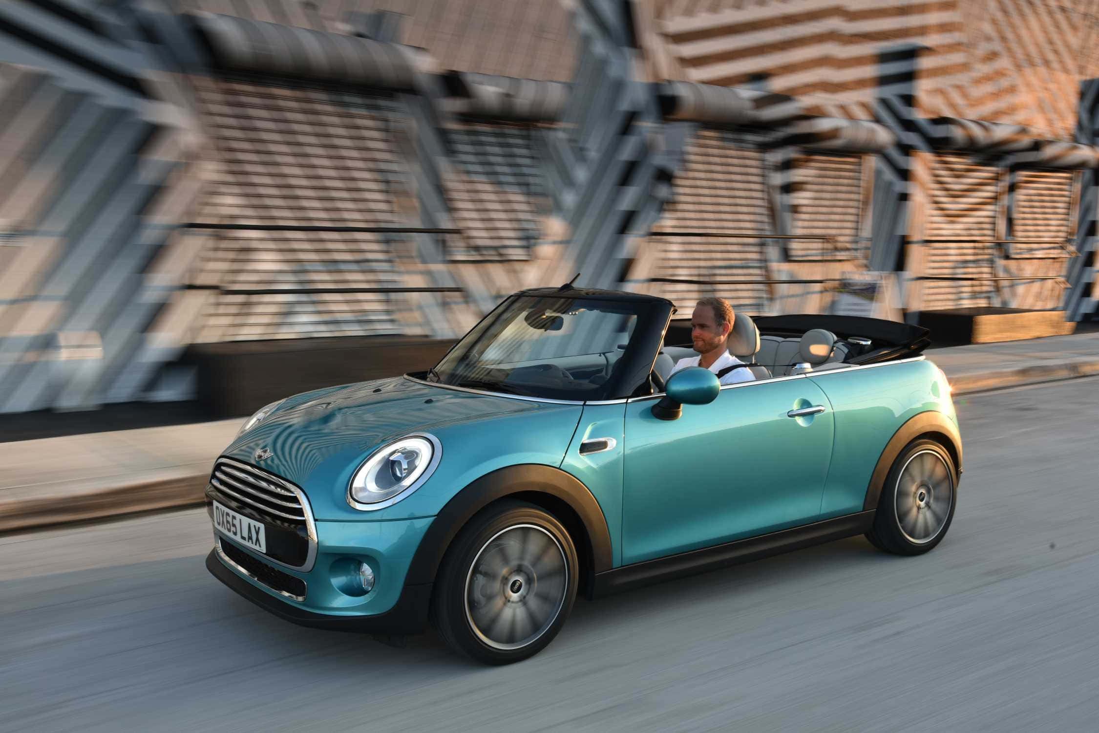 Mini Cooper Convertible 桌布 壁紙 — 一輛青綠色的MINI Convertible在現代城市道路上疾馳，一名男子駕駛經過幾何建築。