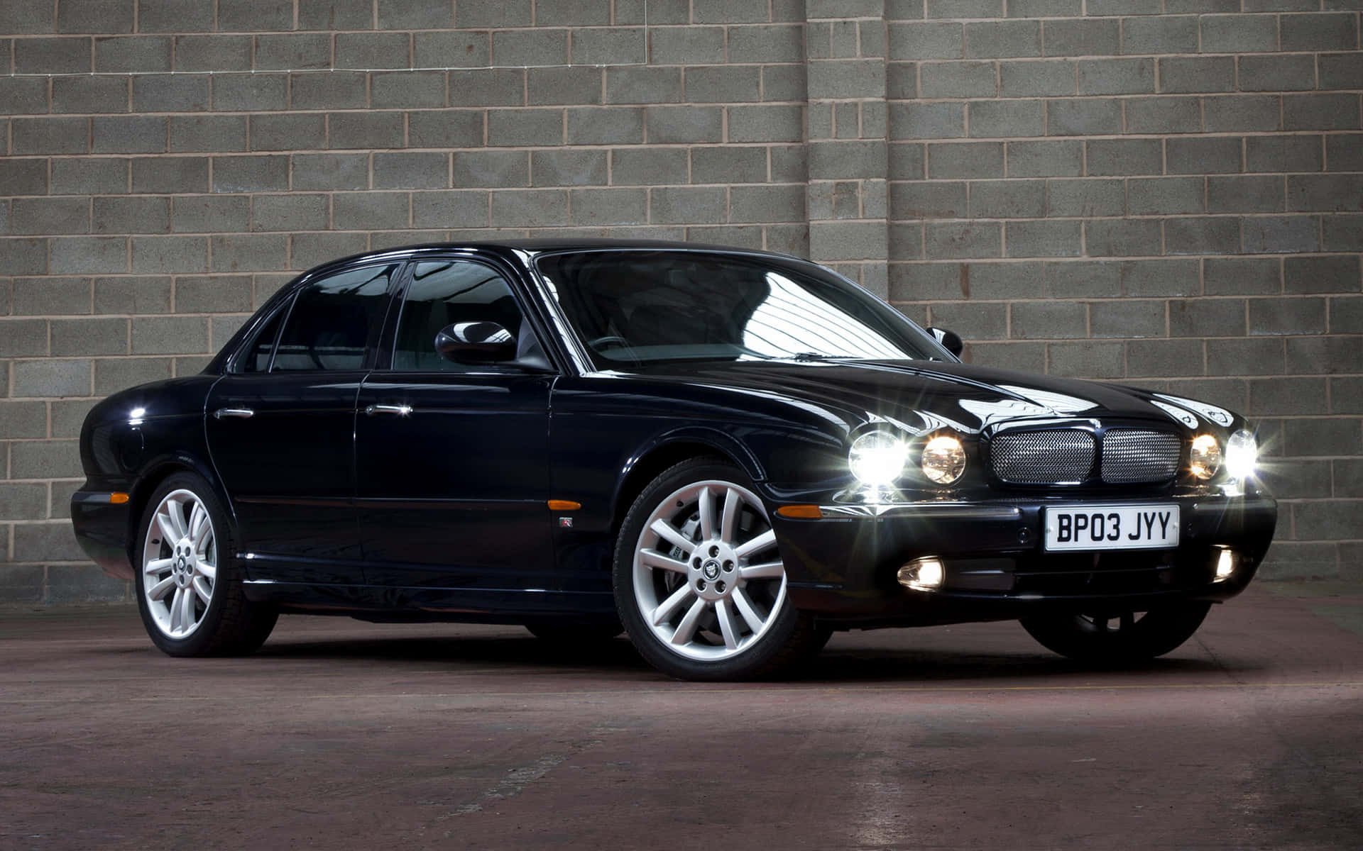 Jaguar Xjr 桌布 壁紙 — 一輛時尚的黑色積架轎車在攝影棚燈光下，以灰色磚牆為背景，亮著車頭燈閃閃發光。