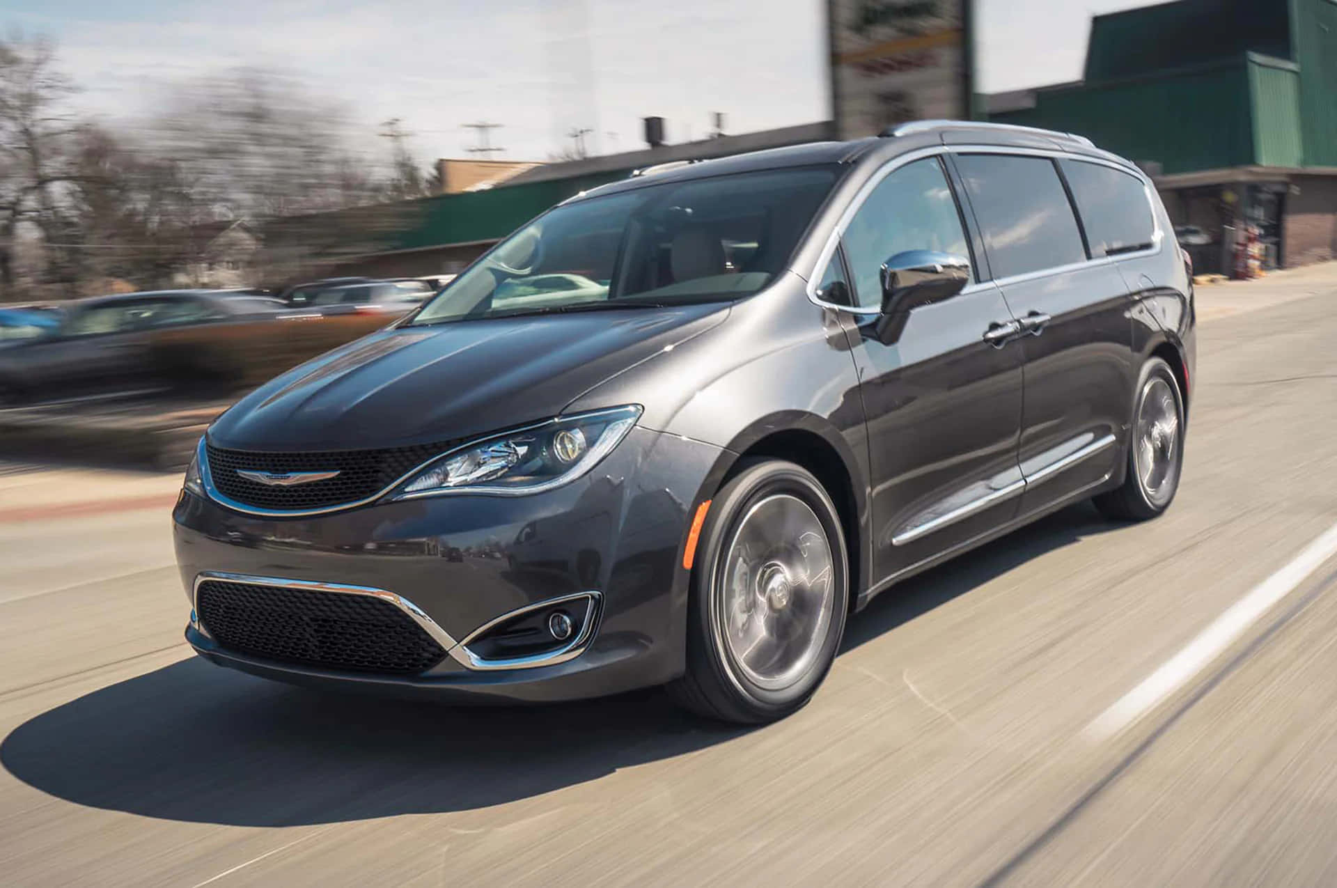 Chrysler Pacifica 桌布 壁紙 — 一輛時尚的灰色小型貨車在陽光明媚的街道上行駛，車輪和背景建築物呈現動態模糊效果。
