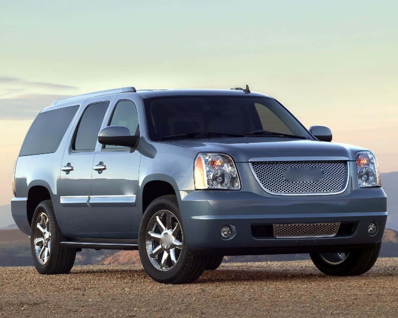 Gmc Yukon 桌布 壁紙 — 一輛時尚的銀色SUV停泊在碎石上，背後是柔和的日落天空。