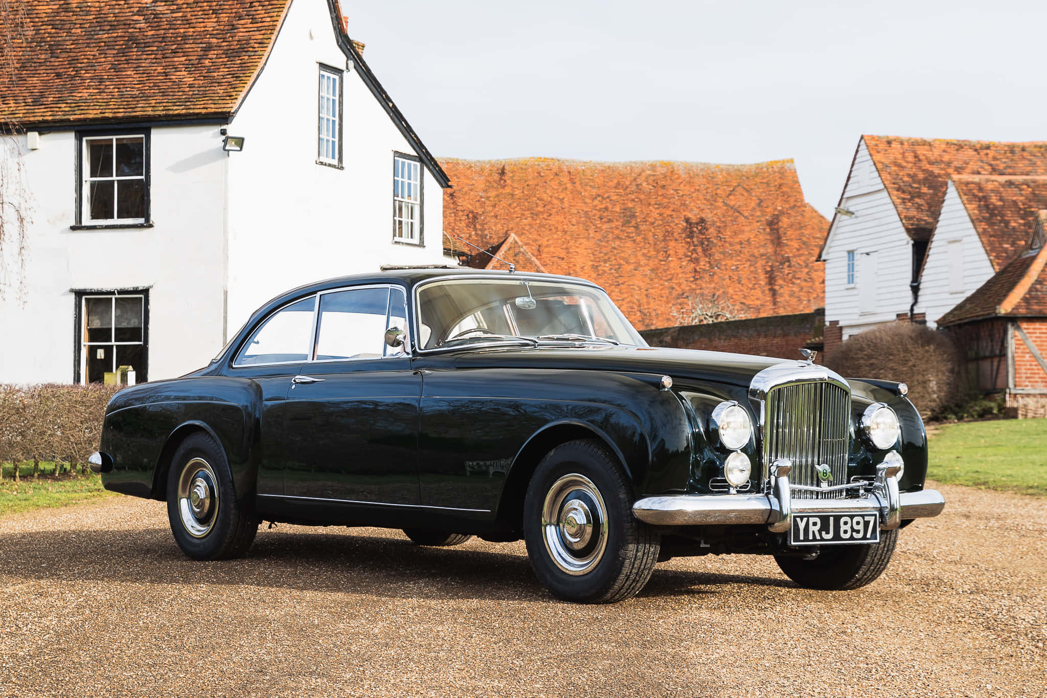 Bentley S2 桌布 壁紙 — 一輛時尚的黑色復古賓利轎車在日光下閃耀，停在碎石路上，前方是白色小屋配陶土屋頂。