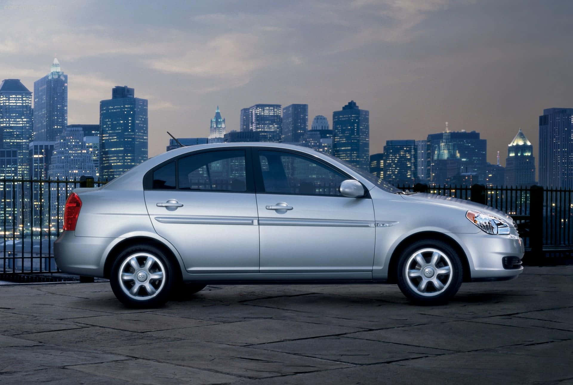 Hyundai Accent 桌布 壁紙 — 一輛銀色轎車停泊在石屋頂上，俯瞰黃昏時分閃爍的城市天際線。