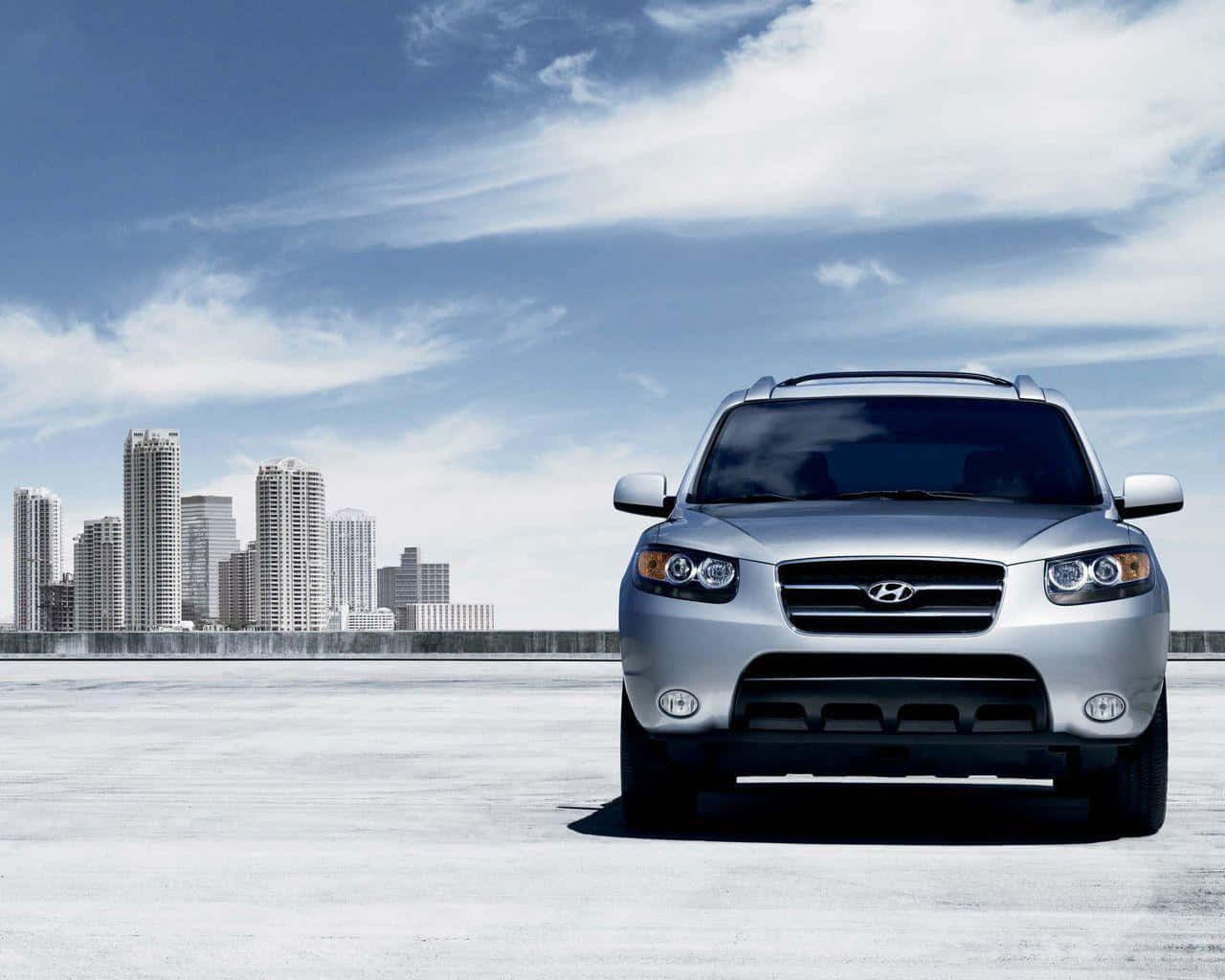 Hyundai Santa Fe 桌布 壁紙 — 一輛銀色現代SUV在混凝土平台上向前，背景是現代城市天際線，天空部分多雲。
