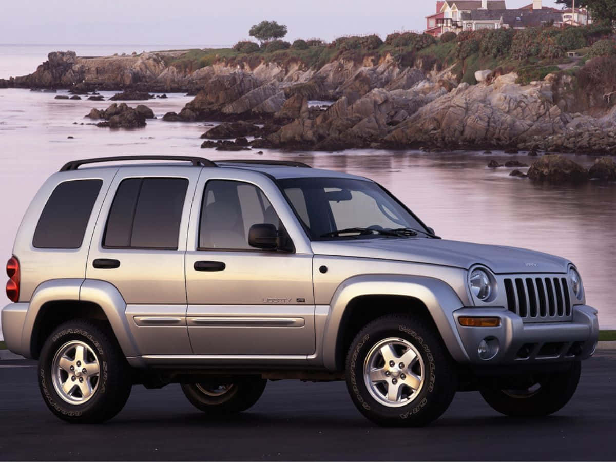 Jeep Liberty 桌布 壁紙 — 銀色Jeep Liberty SUV停泊在黃昏時分岩石海岸線旁的行人路上，懸崖上有房屋。