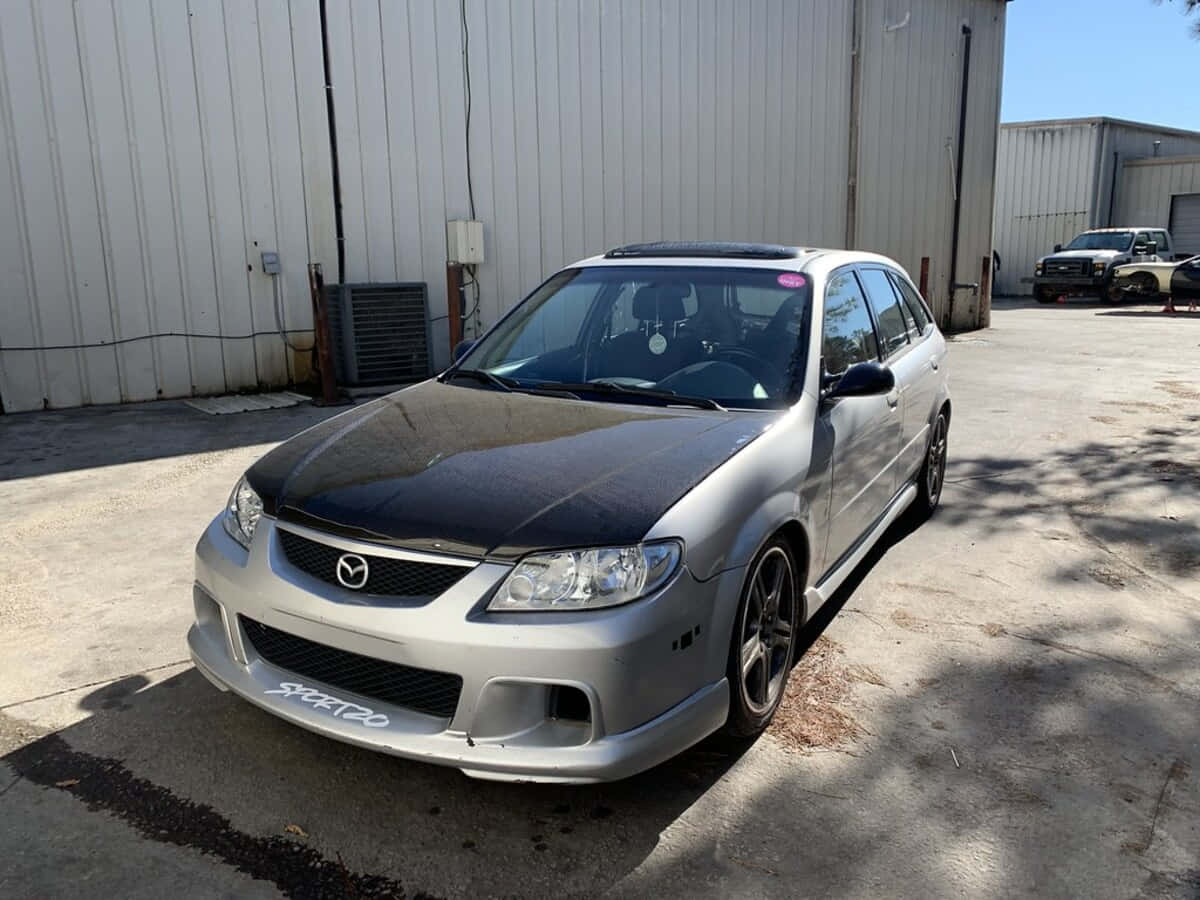 Mazda Protege 桌布 壁紙 — 一輛銀色Mazda 3，黑色引擎蓋和定制輪圈，停在金屬倉庫前，陽光普照。