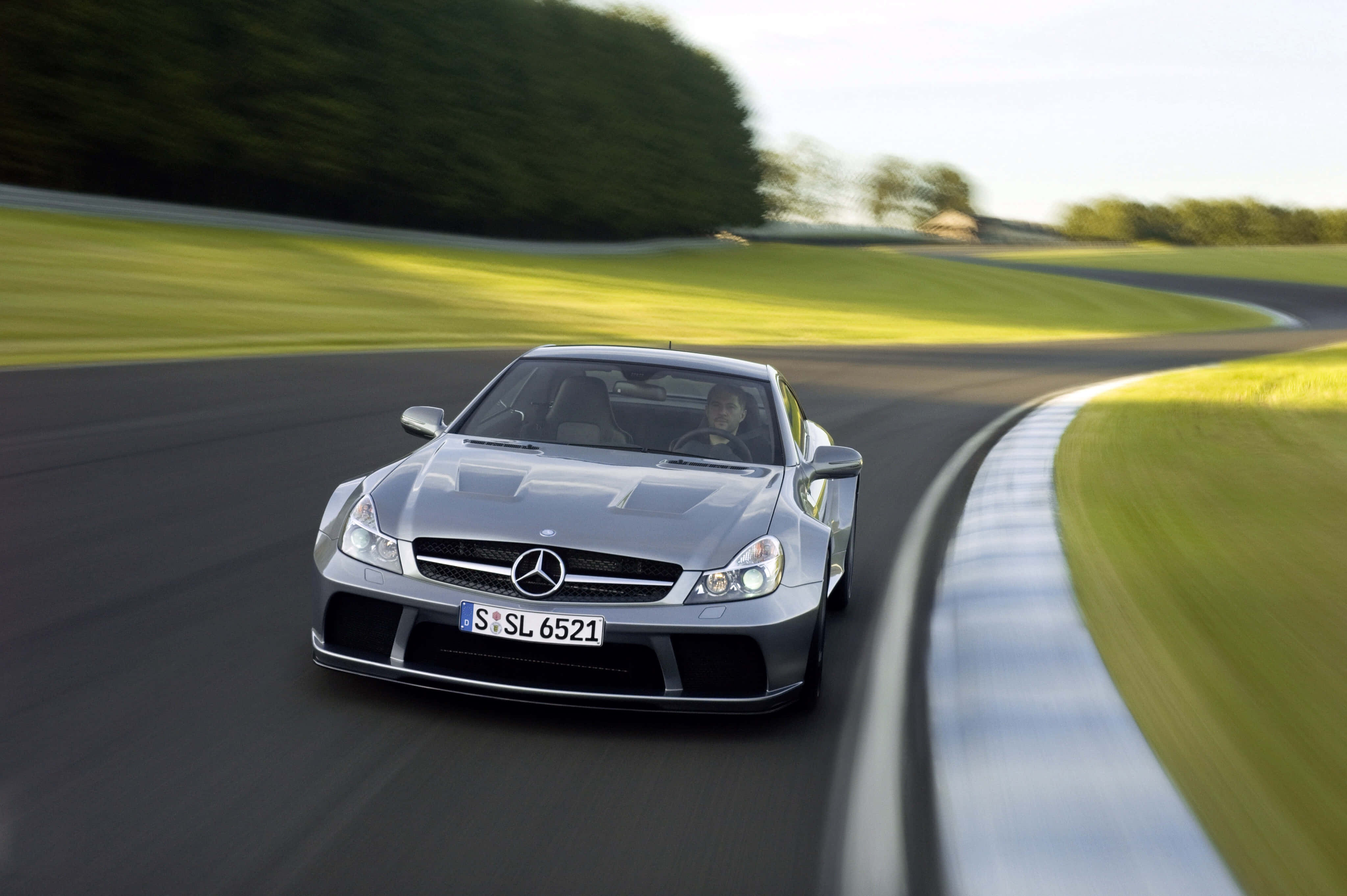 Mercedes Benz Sl-class 桌布 壁紙 — 一輛銀色 Mercedes SL65 AMG 在明亮的日光下，沿著一條被茂密綠草和樹木環繞的彎曲賽道飛馳。