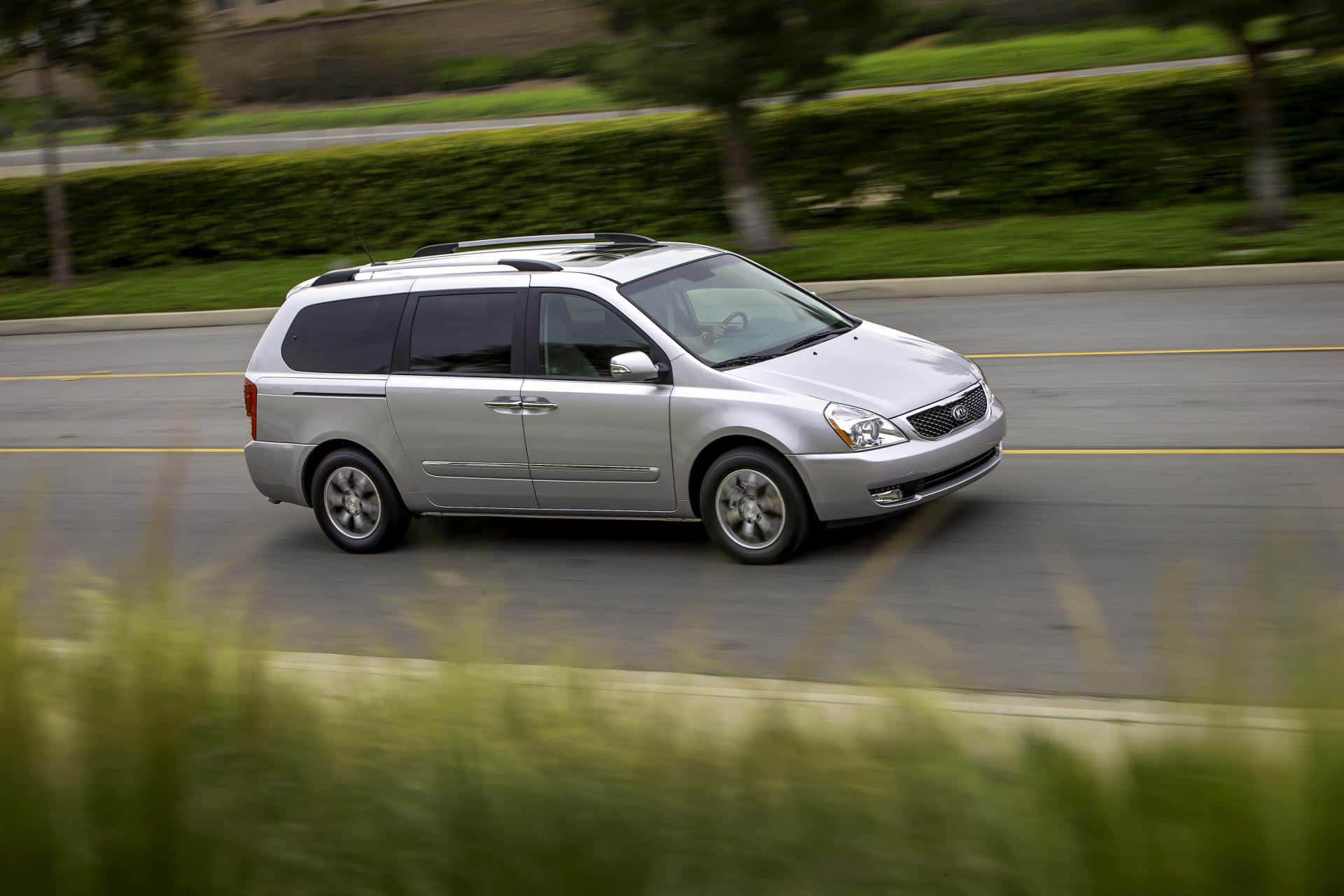 Kia Sedona 桌布 壁紙 — 一輛銀色小型貨車在陽光普照的道路上行駛，道路兩旁是茂密的綠色樹籬和樹木。