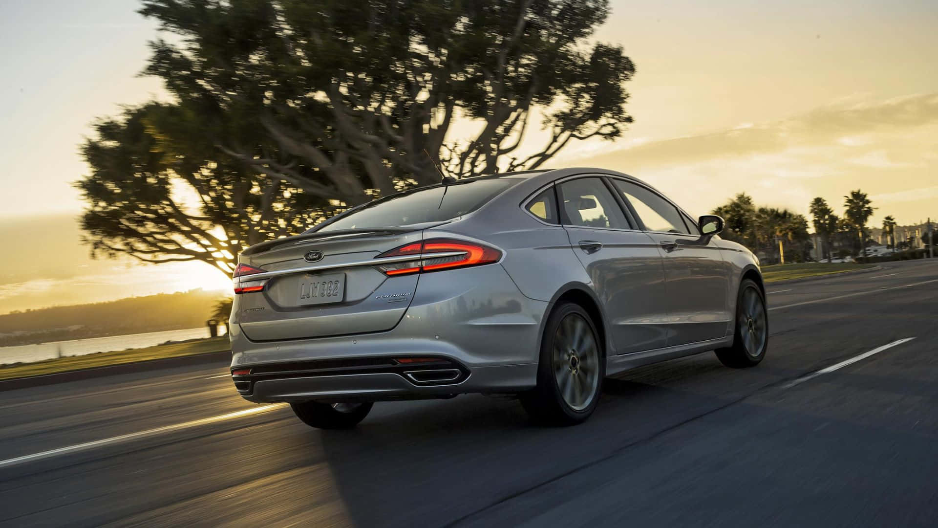 Ford Fusion 桌布 壁紙 — 一輛銀色轎車在黃金時段沿著海岸公路行駛，背景有樹木和水面。
