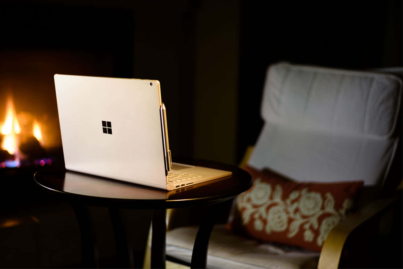  壁紙 — 一部銀色 Microsoft Surface 手提電腦在昏暗的桌子上溫暖地發光，旁邊有噼啪作響的壁爐和一個帶有花卉枕頭的白色扶手椅。