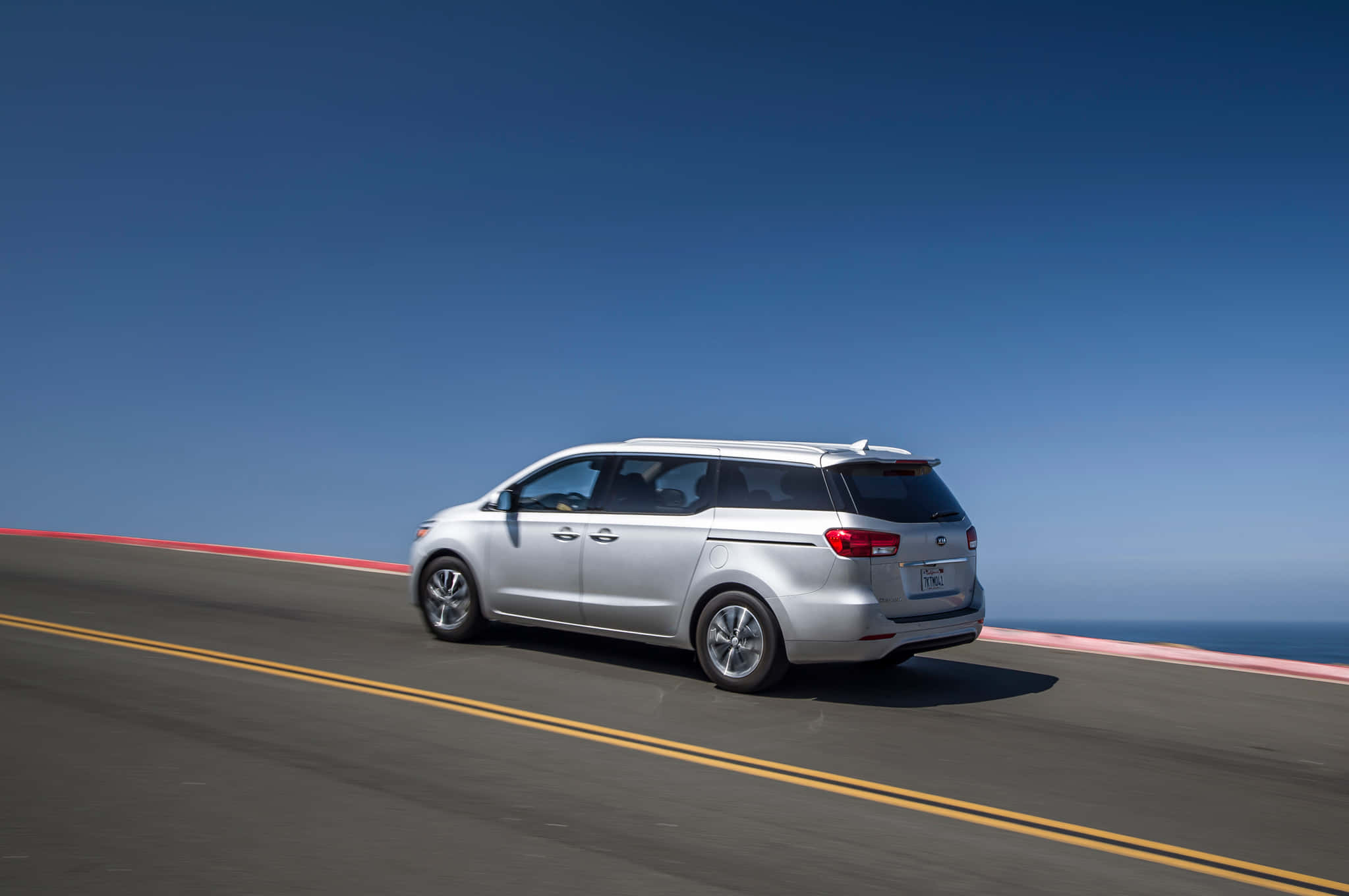 Kia Sedona 桌布 壁紙 — 一輛銀色小型貨車沿著海岸公路行駛，在晴朗的藍天下，海洋地平線盡收眼底。