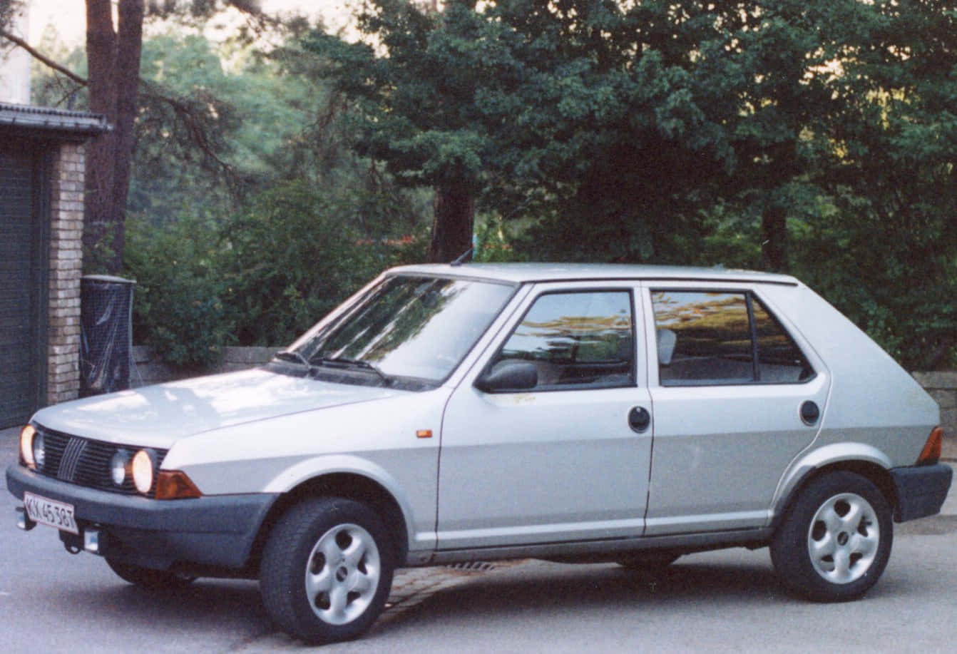 Fiat Ritmo 桌布 壁紙 — 一輛銀色快意烏諾掀背車停泊在鋪砌的車道上，周圍是茂密的綠樹和磚砌建築。