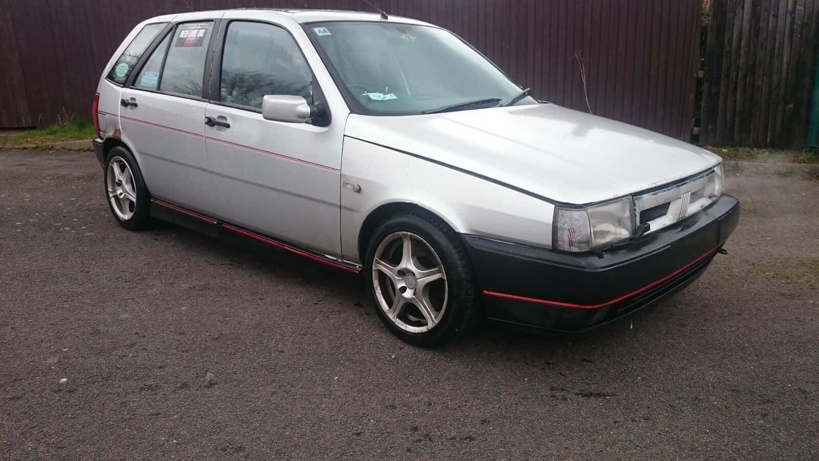 Fiat Tipo 桌布 壁紙 — 一輛銀色掀背車，帶有紅色飾邊和合金輪圈，停在木柵欄附近的瀝青路面上。