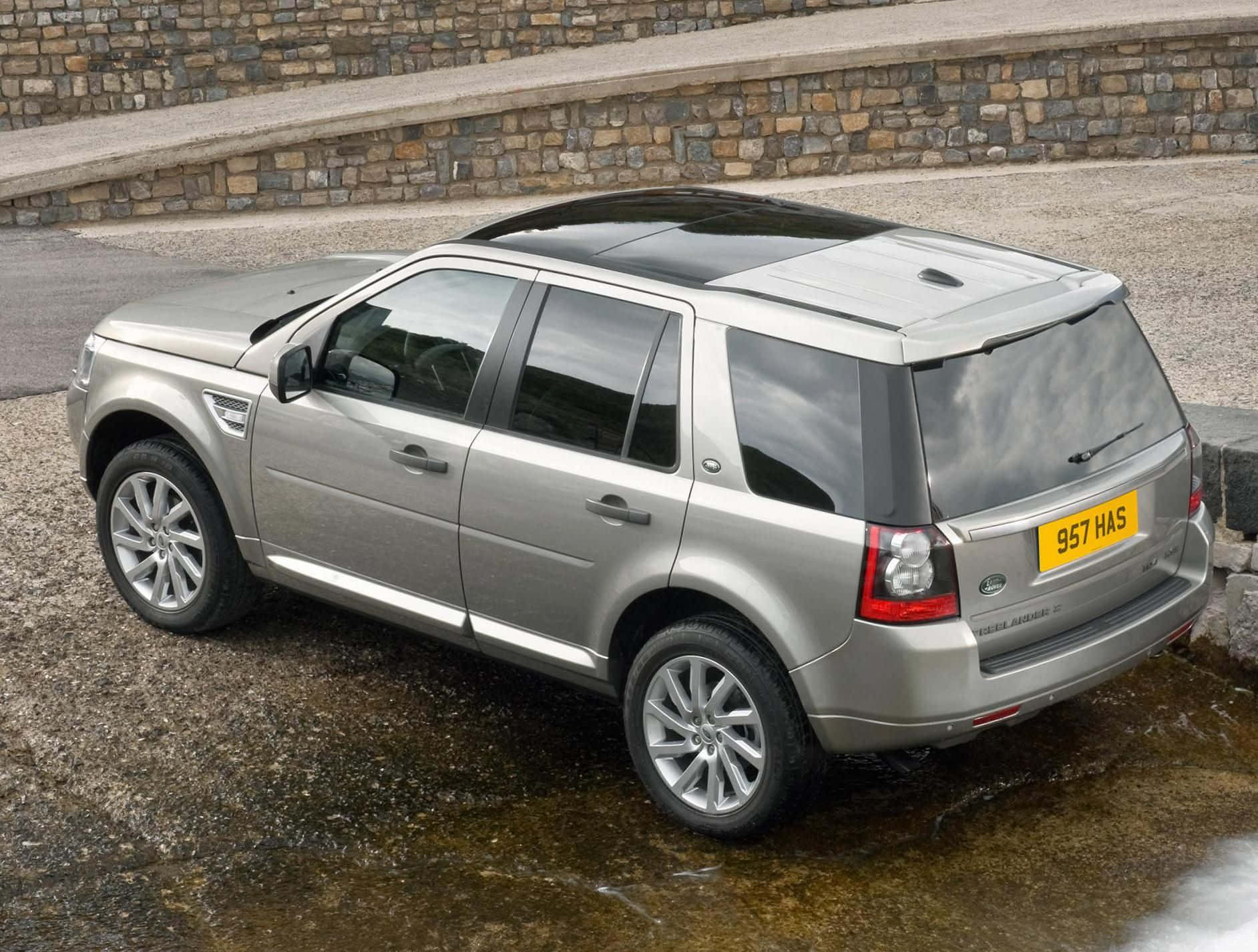 Land Rover Freelander 桌布 壁紙 — 一架銀色 Land Rover Freelander 2 停泊在濕碎石上，旁邊有石牆，輪胎周圍有水積聚。