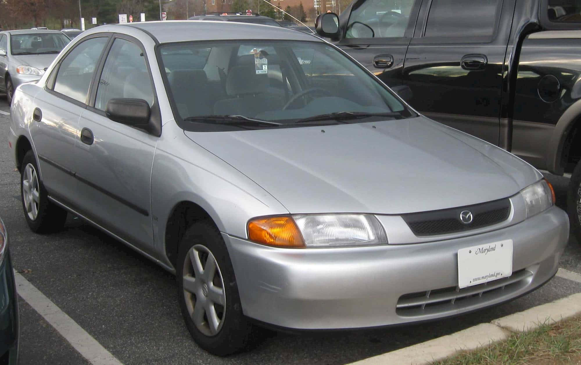 Mazda Protege 桌布 壁紙 — 一輛銀色萬事得房車停泊在戶外停車場，附近有其他車輛。