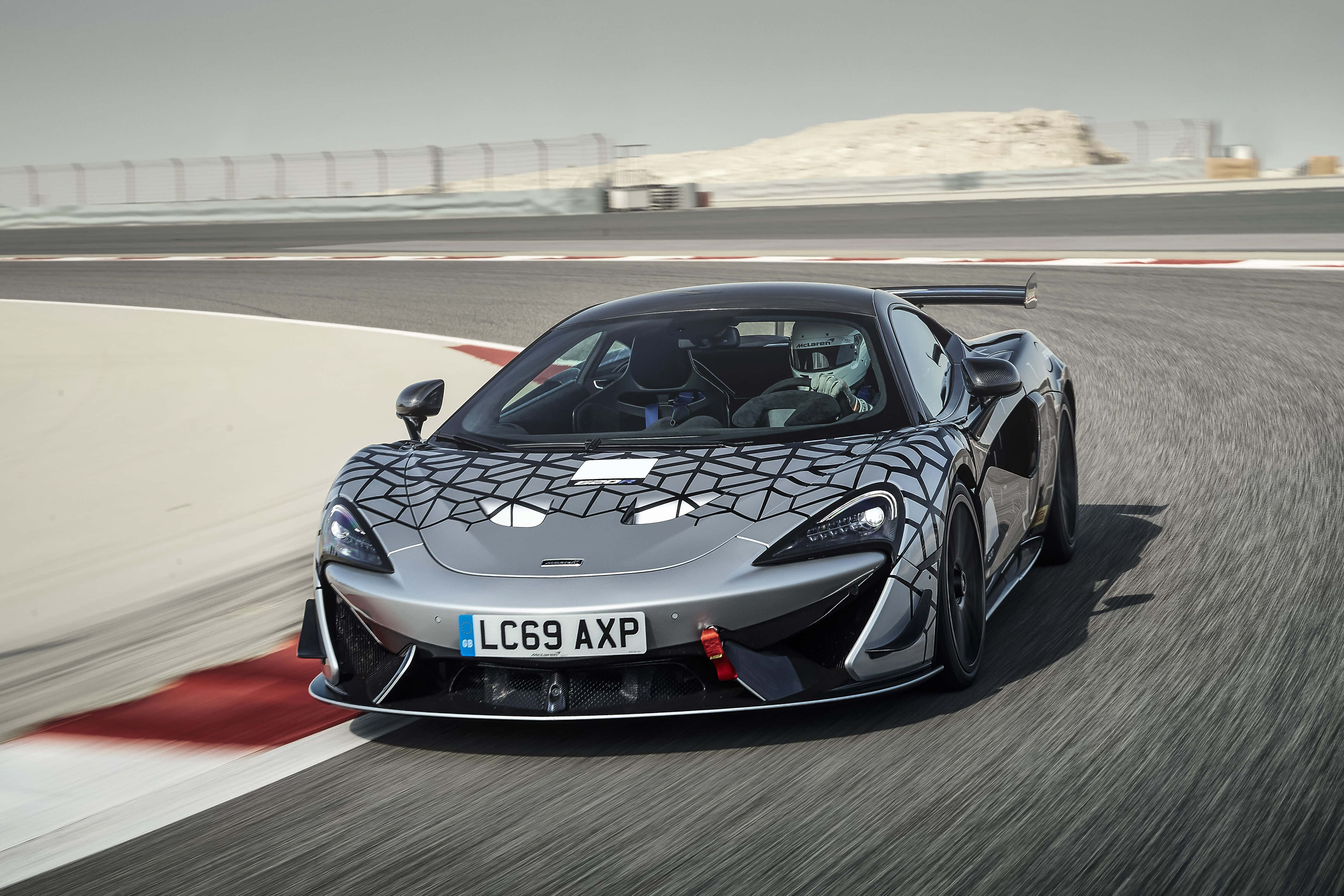 Mclaren 620r 桌布 壁紙 — 一輛銀色McLaren跑車帶有黑色幾何迷彩，在明亮的日光下於賽道彎位疾馳。