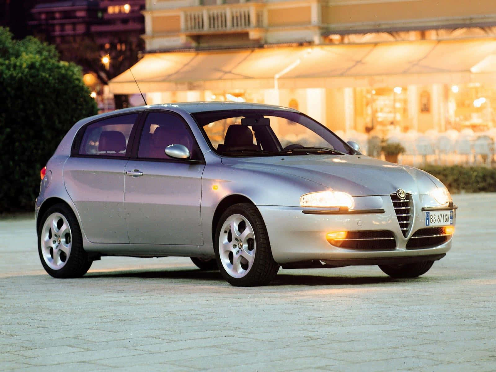 Alfa Romeo 147 桌布 壁紙 — 一輛銀色愛快羅密歐掀背車在溫暖的晚燈下閃閃發光，停在燈火通明的餐廳露台前。