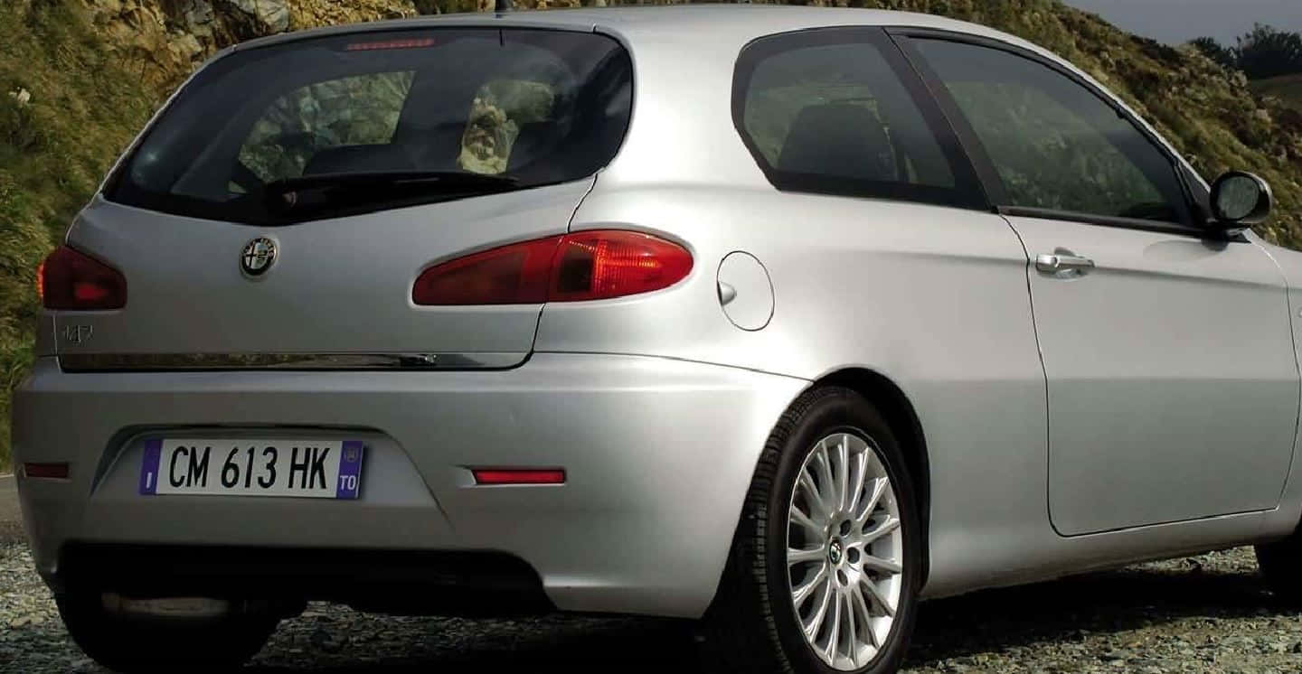 Alfa Romeo 147 桌布 壁紙 — 銀色愛快羅密歐147掀背車停在碎石上，背景係岩石山丘，天色陰沉。