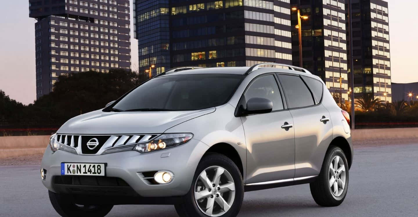 Nissan Murano 桌布 壁紙 — 一輛銀色日產Rogue SUV在黃昏時停泊在現代玻璃摩天大樓前，窗戶亮著燈。