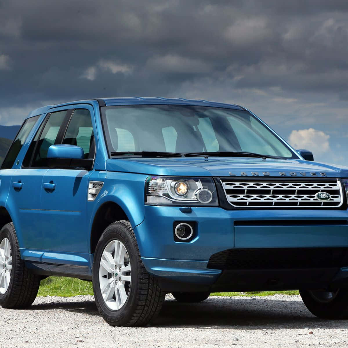 Land Rover Freelander 桌布 壁紙 — 一輛充滿活力的藍色 Land Rover SUV 停在碎石路上，背景是戲劇性的多雲天空和遠處的山脈。