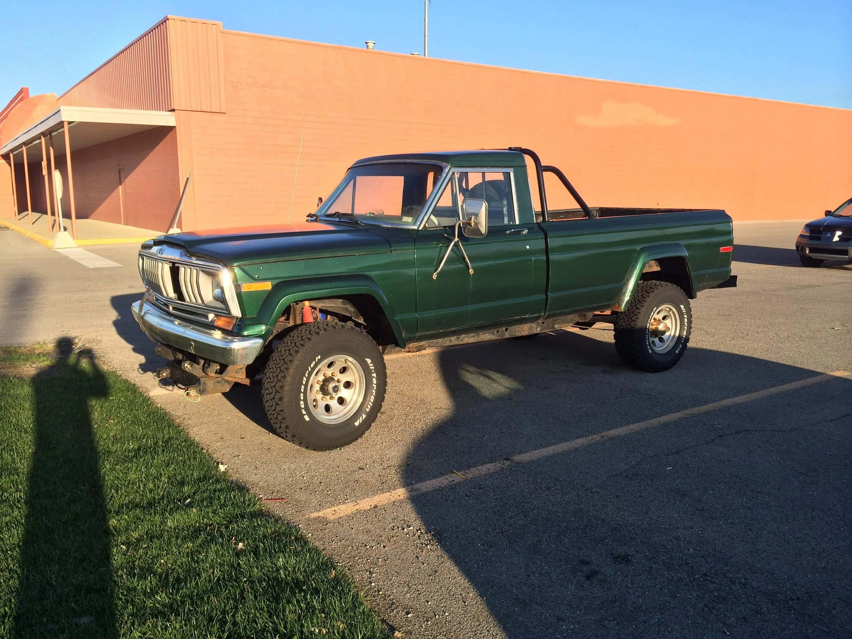 Jeep Comanche 桌布 壁紙 — 一架升高了的綠色經典皮卡車停泊在一個停車場，旁邊是一棟米色建築，天空晴朗蔚藍。