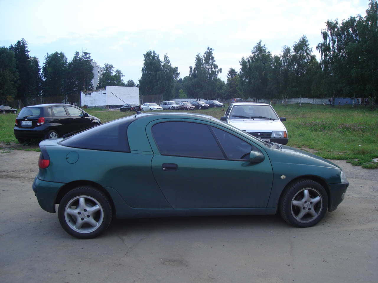 Opel Tigra 桌布 壁紙 — 一架青綠色雙門轎跑車停泊在碎石地上，背景有樹木和其他車輛，天空多雲。