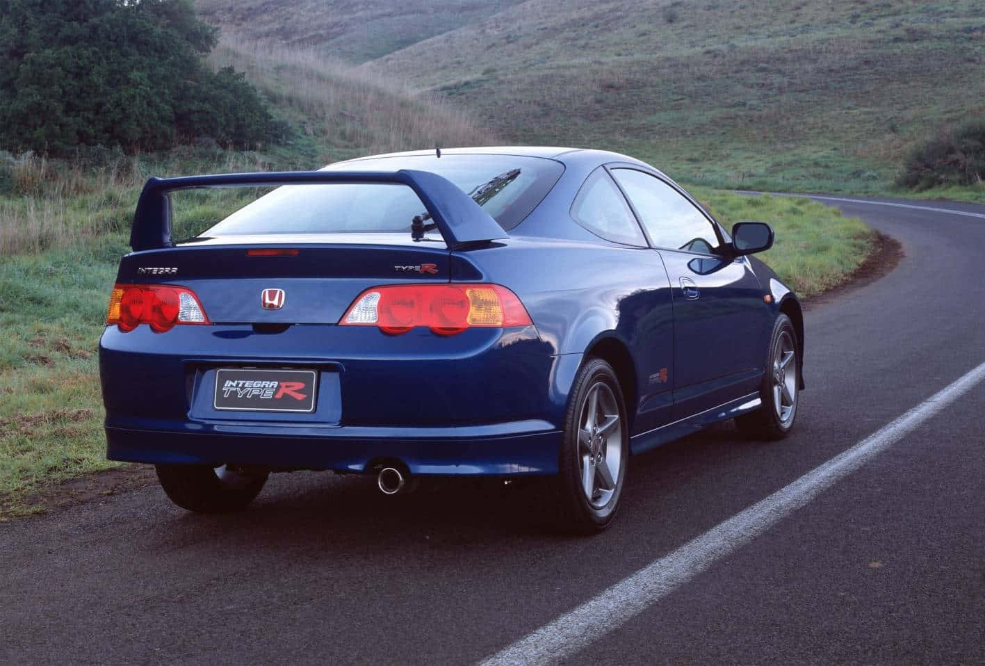 Honda Integra 桌布 壁紙 — 一架流線型的藍色本田Integra Type R轎跑車沿著一條蜿蜒的鄉村道路行駛，兩旁是長滿青草的山丘。
