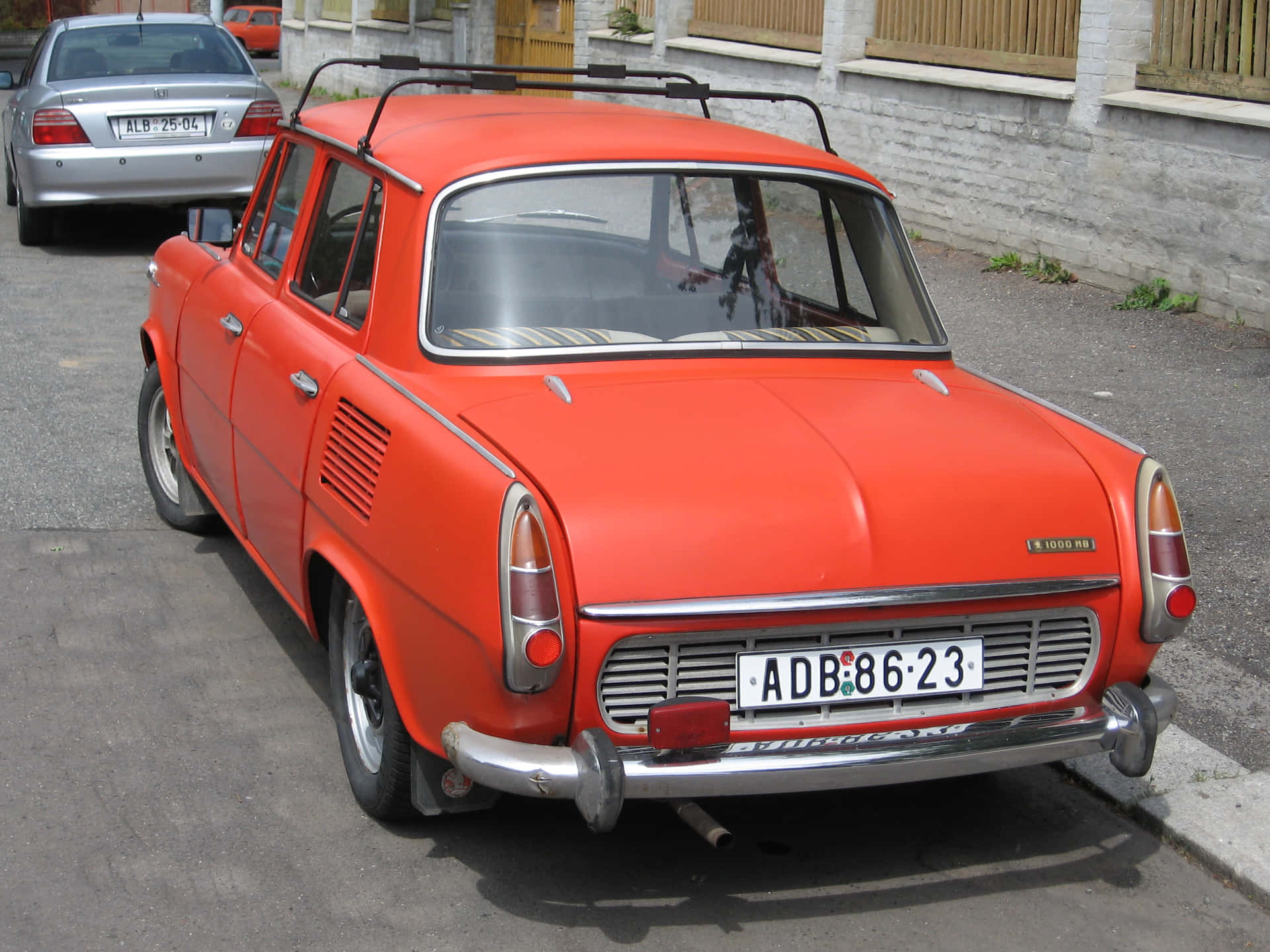  壁紙 — 一輛復古橙色 Trabant 汽車停泊在鋪砌的街道上，旁邊是白色磚牆。