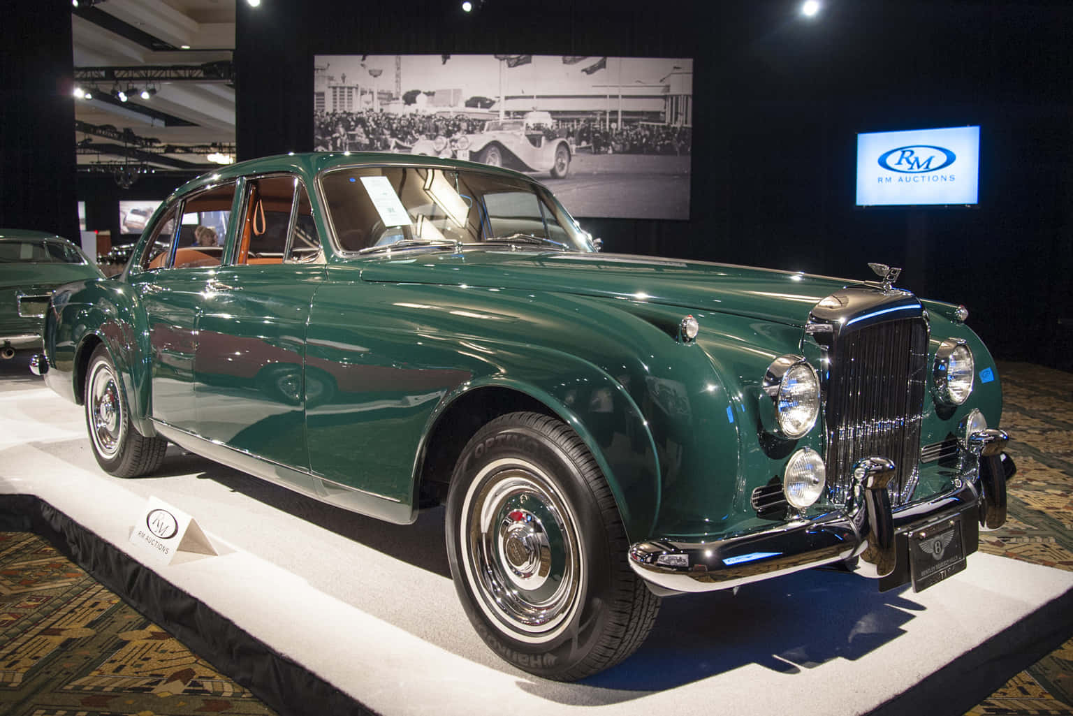 Bentley S2 桌布 壁紙 — 一輛光澤的綠色復古積架轎車在室內汽車拍賣會上，在聚光燈下於白色展示平台上閃閃發光。