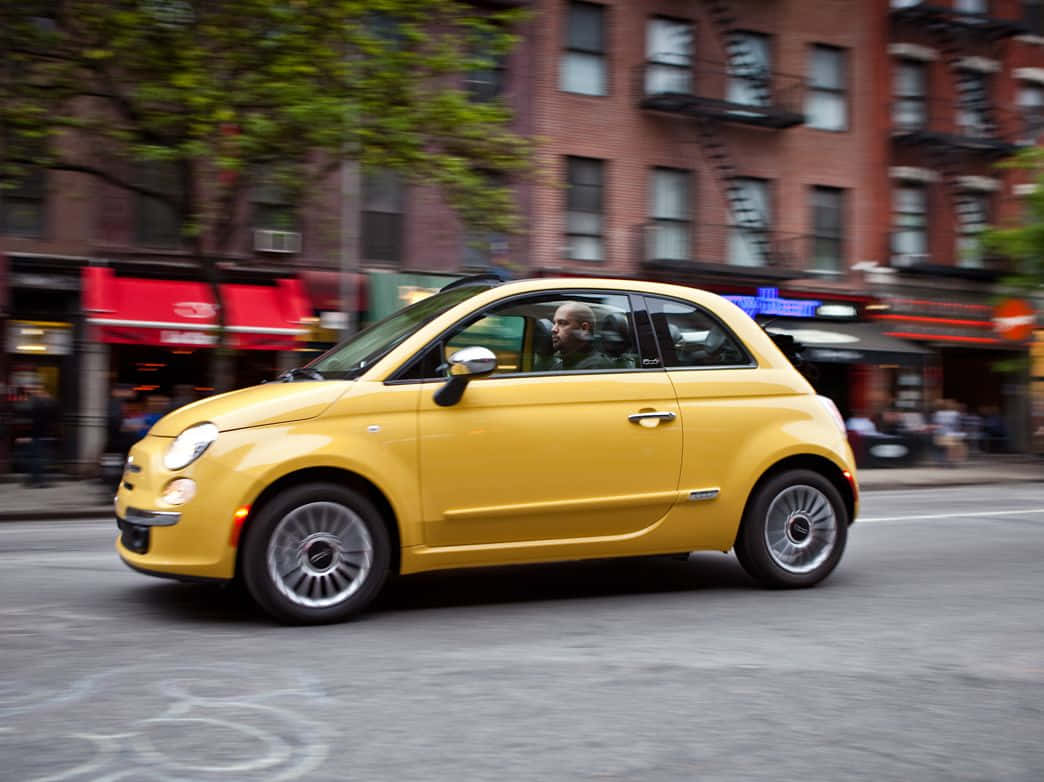 Fiat 500 桌布 壁紙 — 一輛亮黃色的Fiat 500在繁忙的城市街道上行駛，背景的建築物和樹木呈現動態模糊效果。