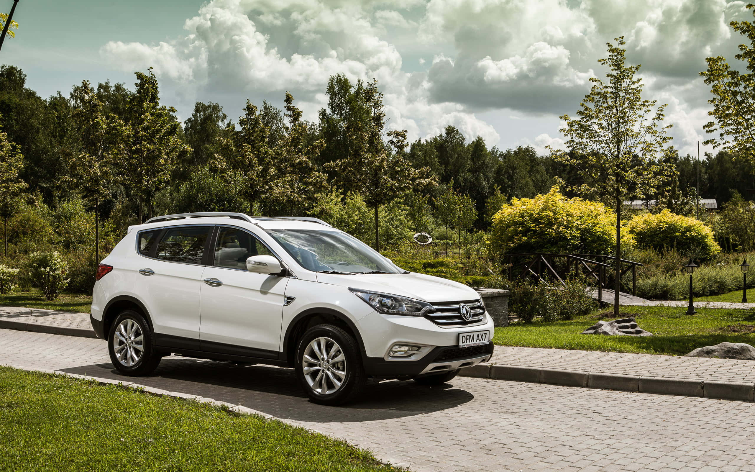 Dongfeng 桌布 壁紙 — 一輛白色SUV停泊在鋪砌小徑上，旁邊有綠草和樹木，天空多雲。