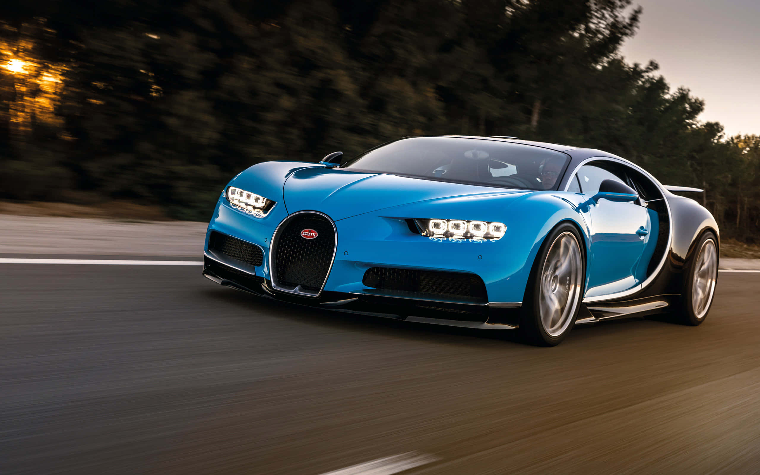 Bugatti Chiron 桌布 壁紙 — 一輛充滿活力的藍色Bugatti Chiron在日落時分沿著蜿蜒的道路疾馳，背景是模糊的樹木。