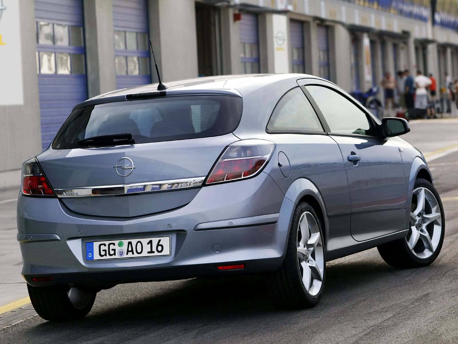 Opel Astra 桌布 壁紙 — 一輛銀色歐寶雅特GTC停泊在鋪砌的停車場，背景有工業建築物和人群。