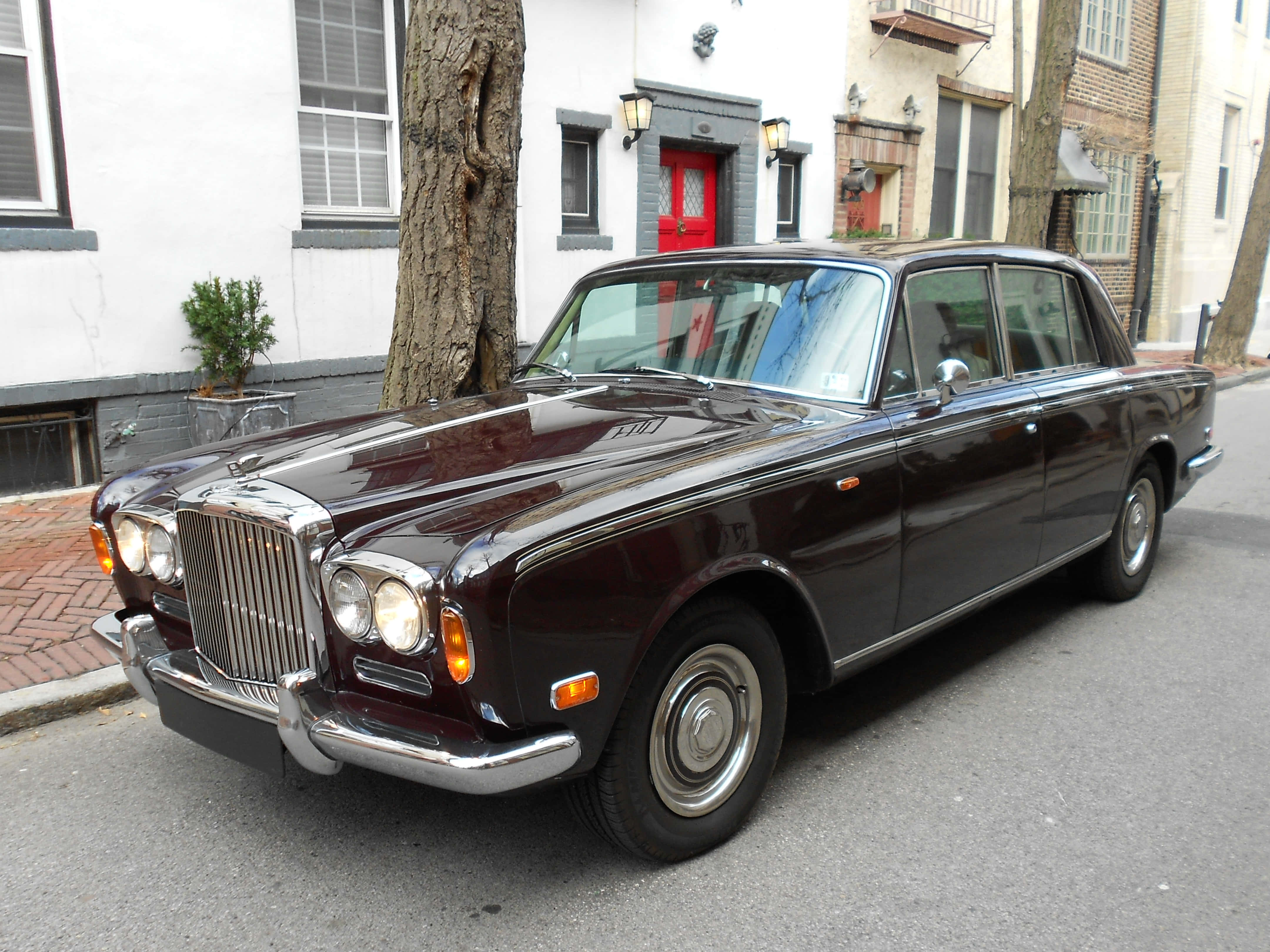 Bentley T1 桌布 壁紙 — 一輛亮面酒紅色的賓利S1轎車停在磚砌街道上，旁邊有一棵樹和一座帶紅門的白色建築。
