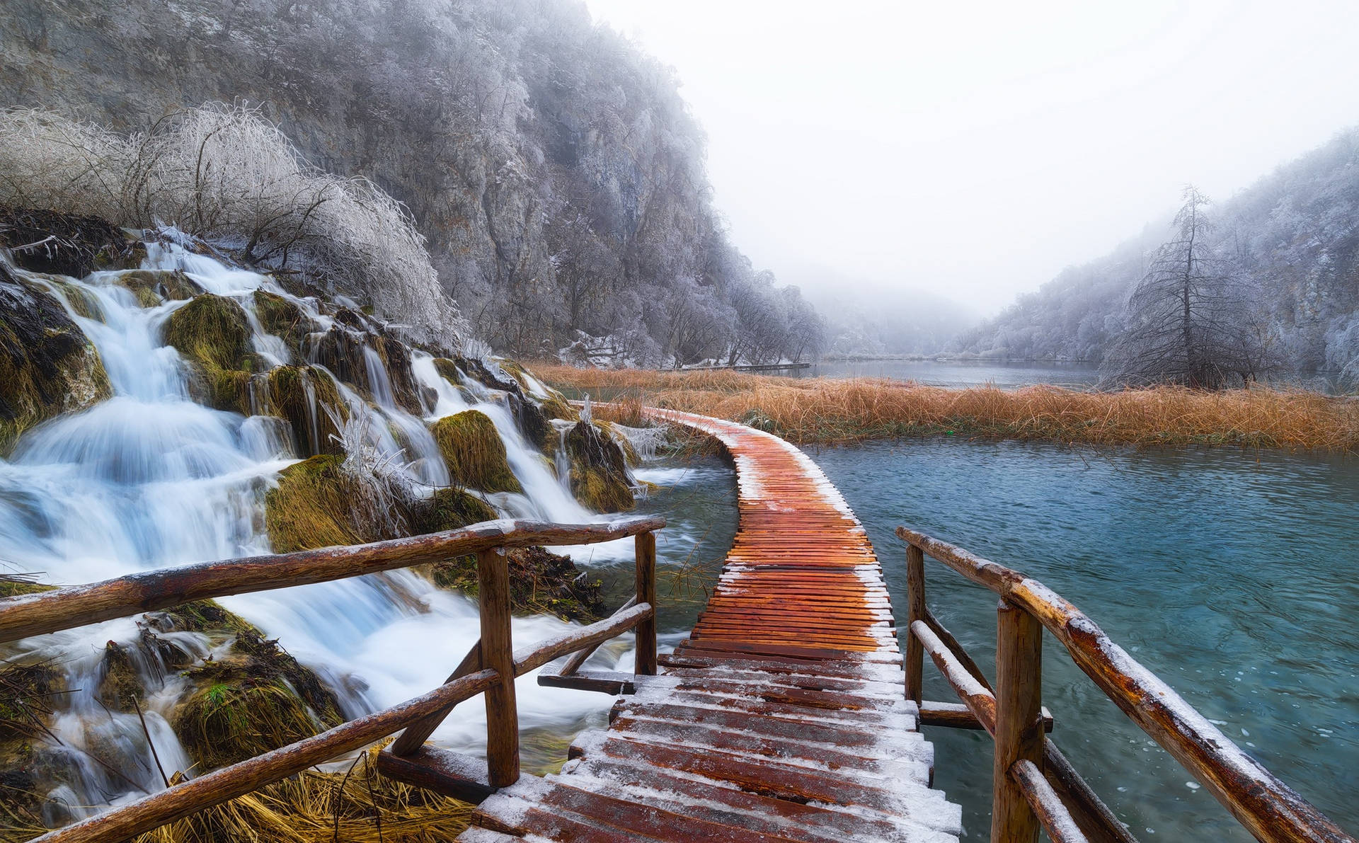 冬季中，被雪覆蓋的木棧道旁邊有流動的瀑布和藍色的湖泊。