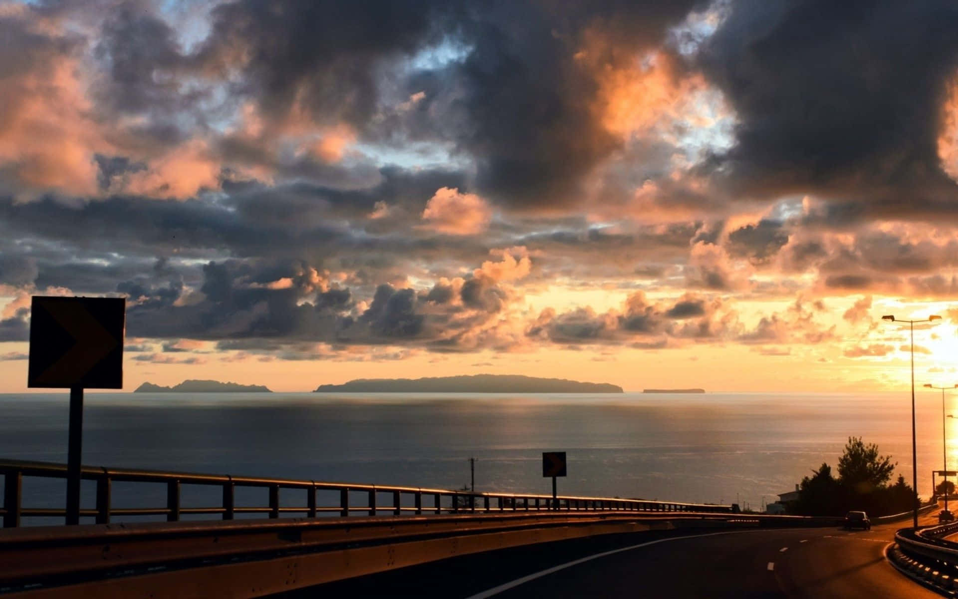 Coastal Sunset 桌布 壁紙 — 一條蜿蜒的沿海高速公路彎向日落，熾熱的橙色雲彩在平靜的海面和遠處的島嶼上空發光。