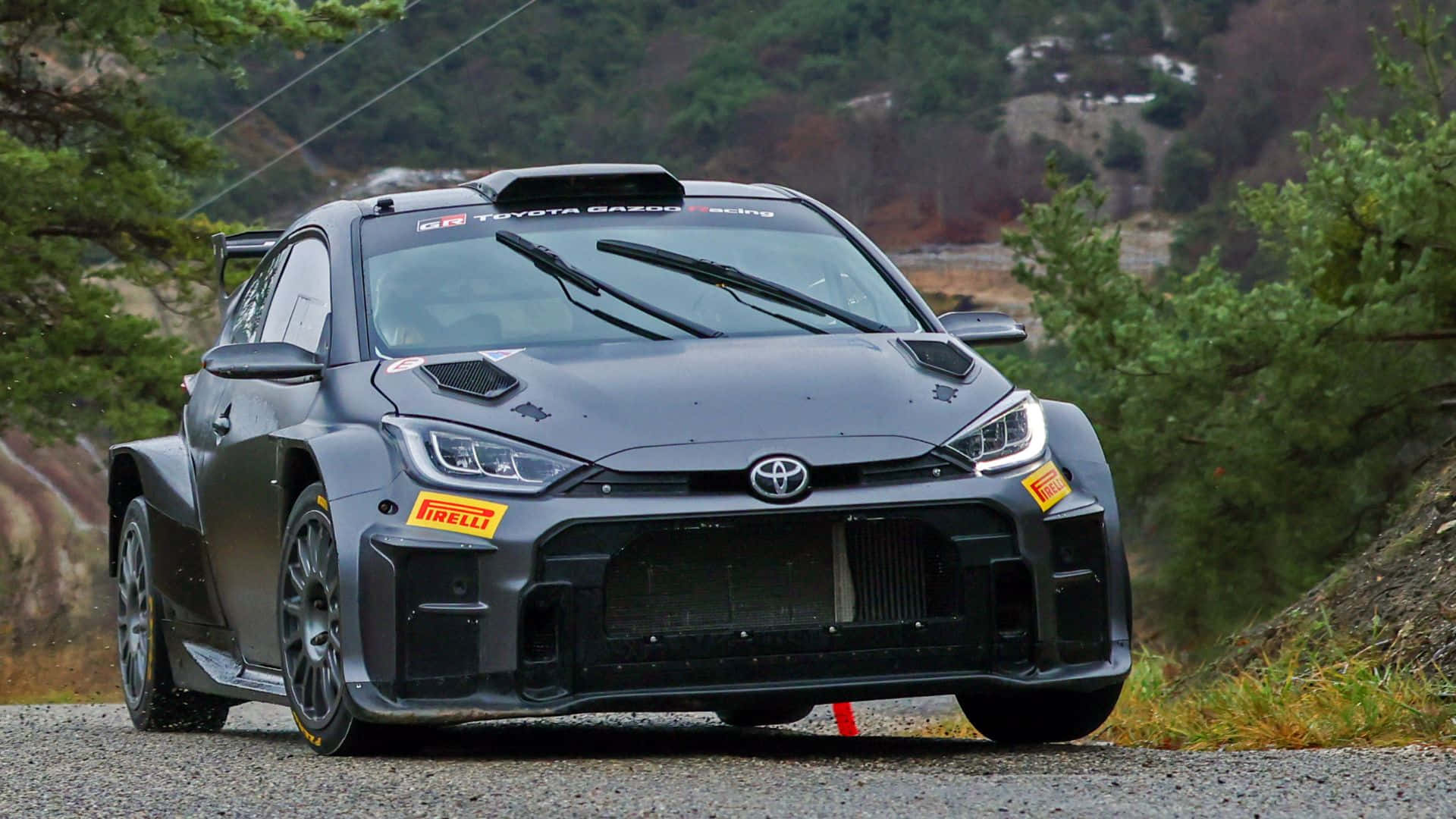  壁紙 — 一輛啞光灰色的豐田GR雅力士Rally1賽車在樹木和山丘背景下的碎石森林道路上飛馳。