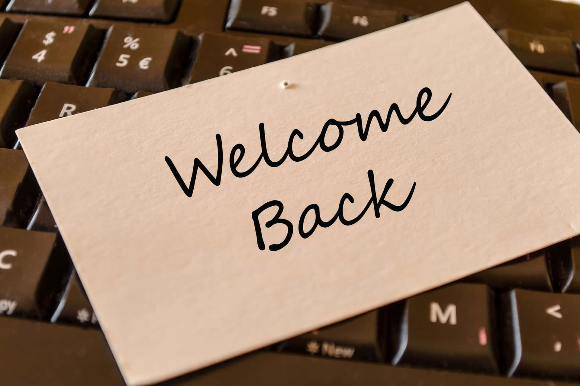  壁紙 — 一張手寫的「Welcome Back」便條放在一個深色電腦鍵盤上，鍵盤上的按鍵和符號清晰可見。