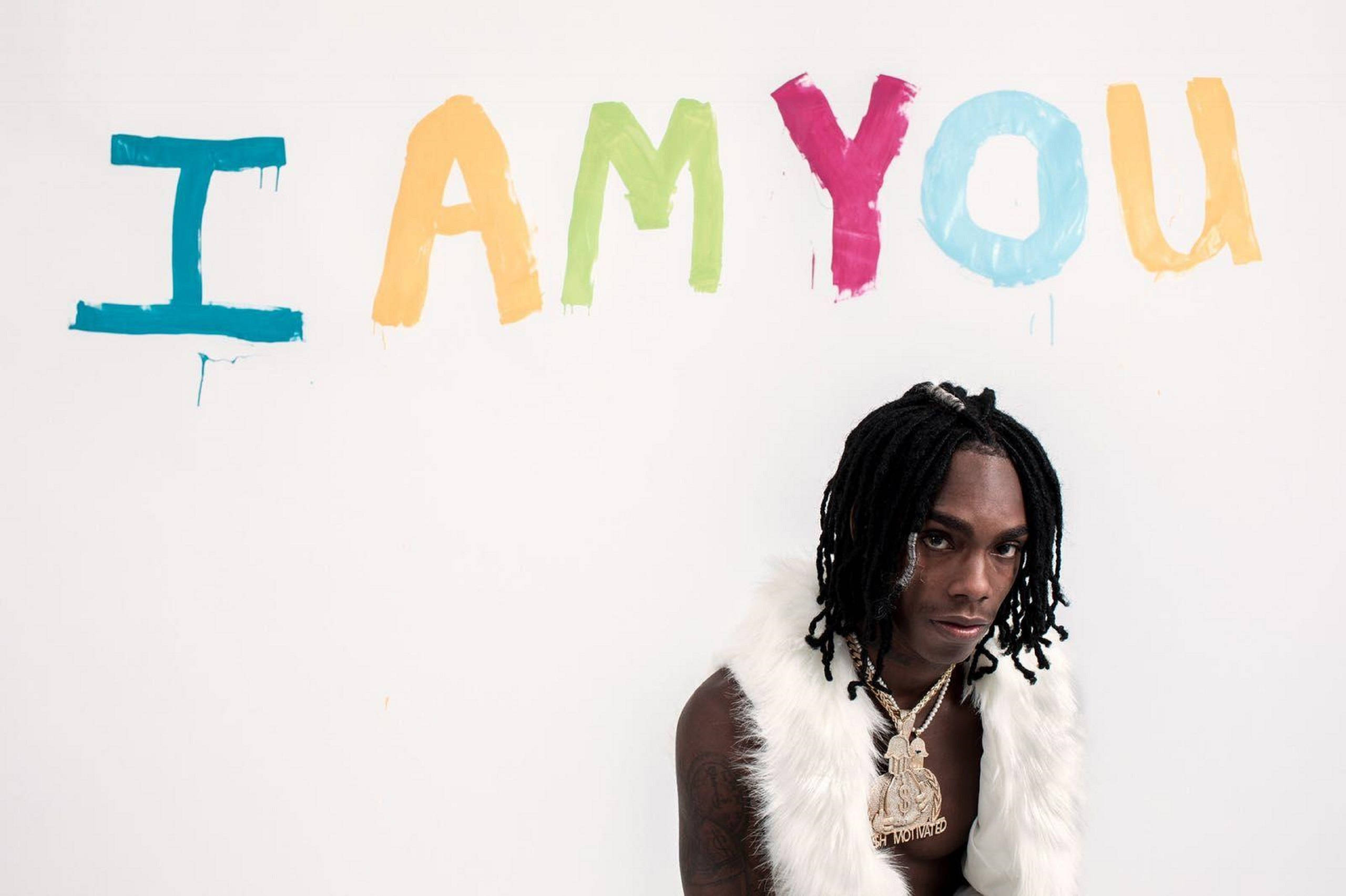 YNW Melly I Am You 迈阿密 壁紙 — 一位留著辮子、身穿白色毛背心的年輕男子站在一幅以粗獷筆觸塗上色彩繽紛的「I AM YOU」字句的牆前。