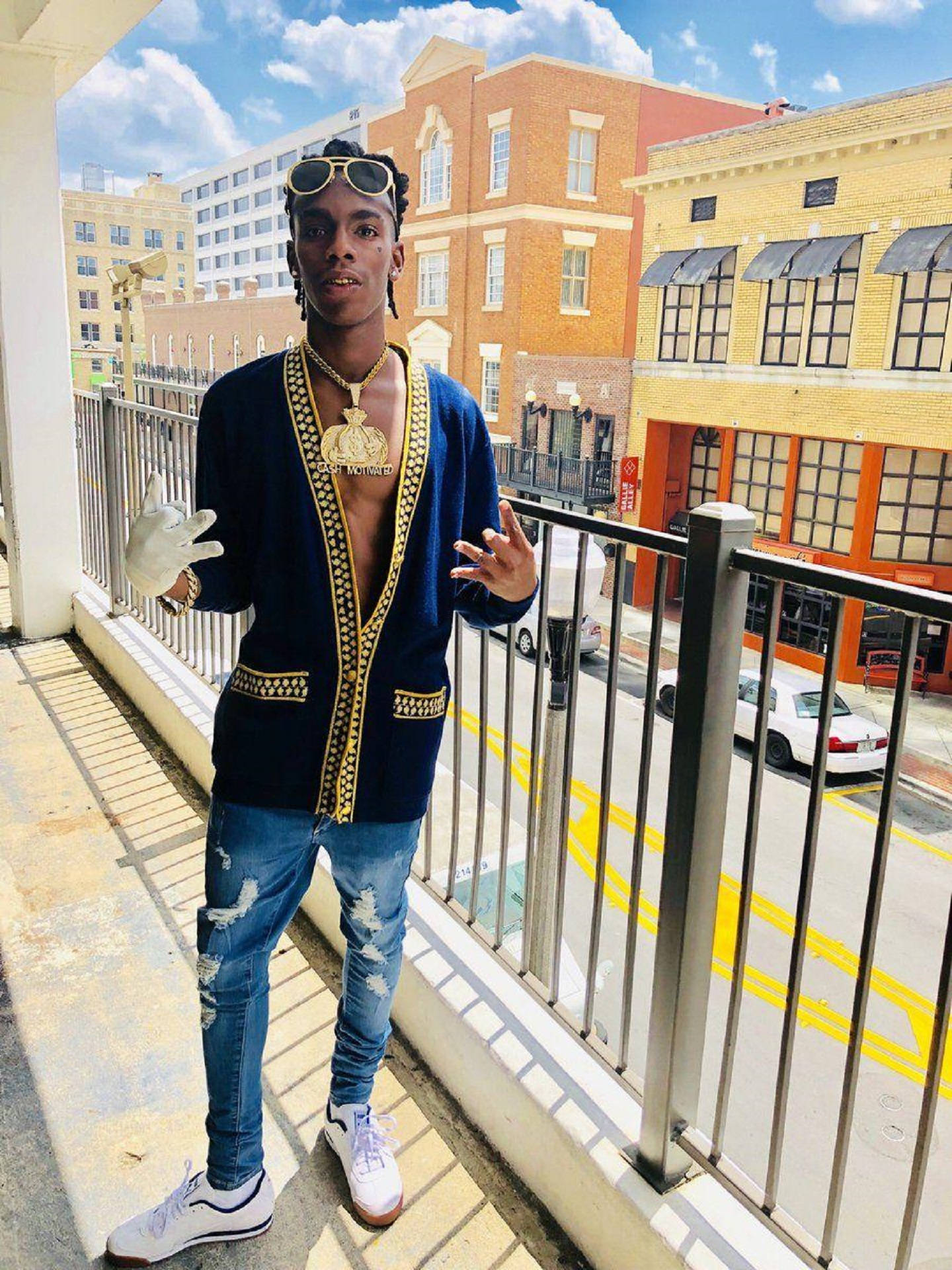YNW Melly《In Town》街景寫真 壁紙 — 一位年輕男子身穿金色刺繡藍色開襟衫和破洞牛仔褲，在陽光明媚的城市陽台上自信擺姿勢，身後是城市建築。