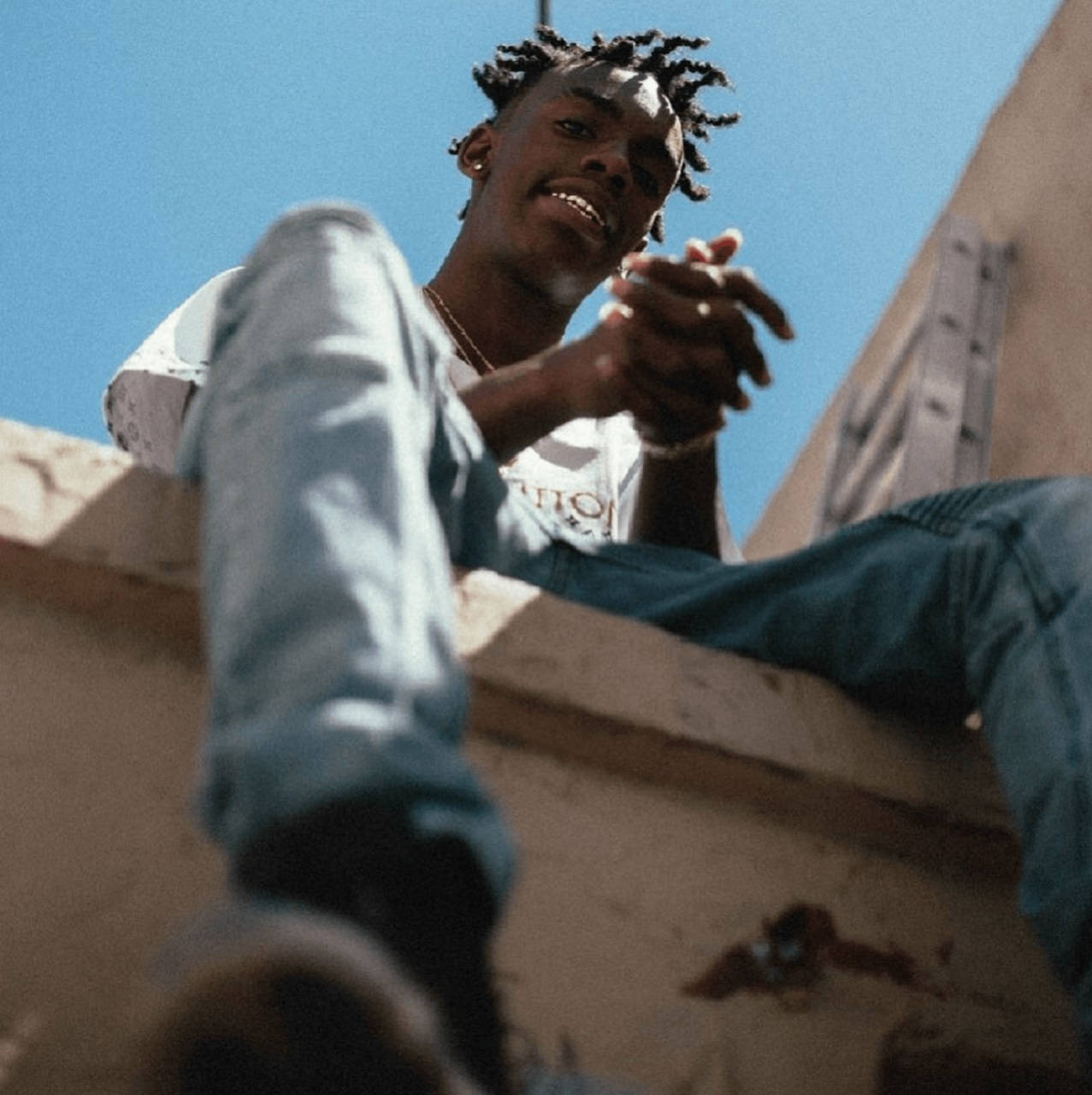 YNW Melly《Slang That Iron》屋頂低角度美學照 壁紙 — 一位留著辮子的年輕男子在晴朗的藍天下從屋頂邊緣微笑俯視。