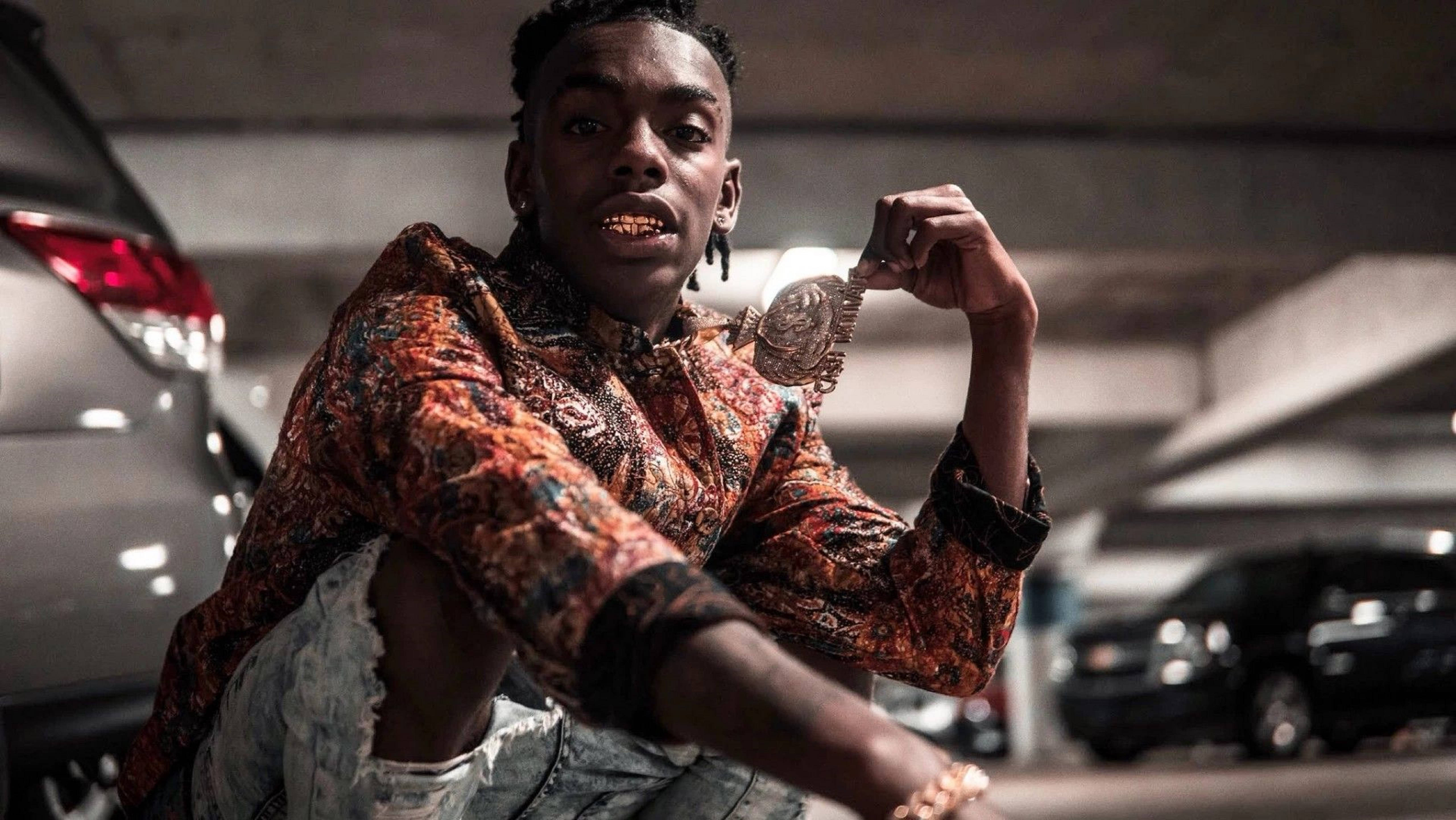 YNW Melly《Slang That Iron》專輯封面 壁紙 — 一位身穿鮮豔圖案襯衫的年輕男子在光線昏暗的停車場自信地擺姿勢，手持一條大型裝飾鏈條。