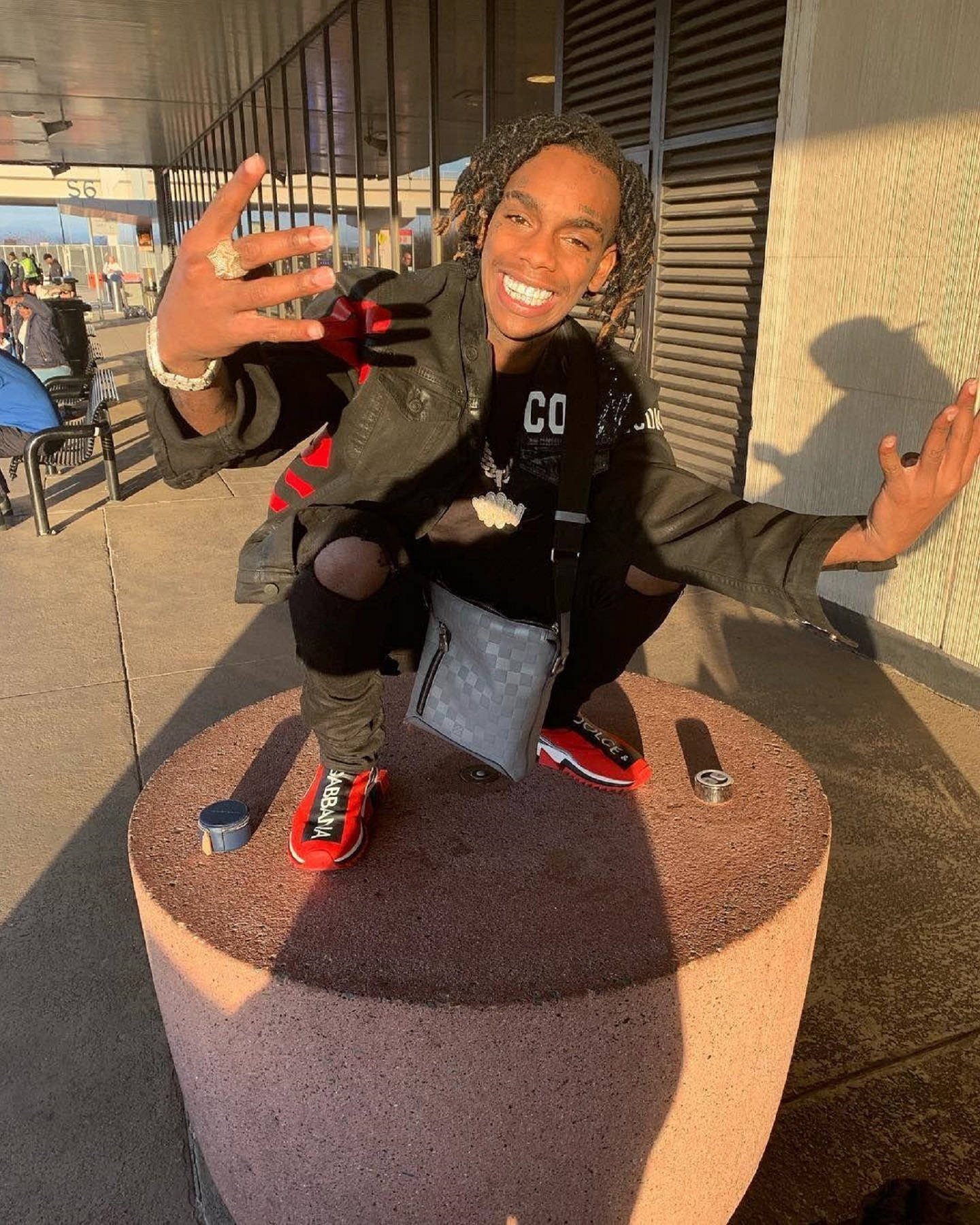 YNW Melly《The Melly Way》宣傳照 壁紙 — 一位微笑的年輕男子留著辮子，蹲在戶外的混凝土柱上，穿著紅色運動鞋和黑色外套，雙手做手勢。