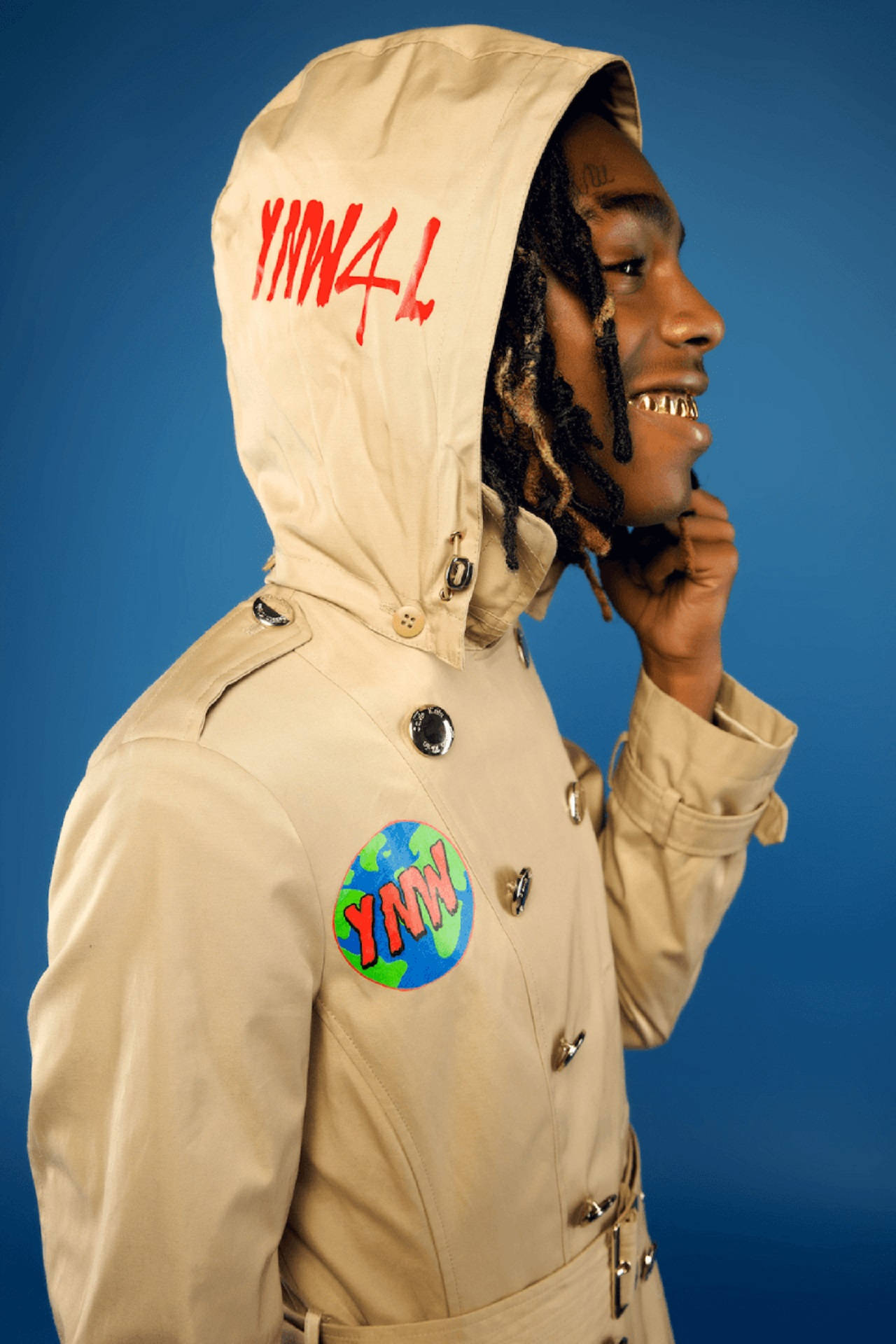 YNW Melly《We All Shine》巡迴海報 壁紙 — 一名身穿米色風衣、帽子上有「YNN4L」塗鴉和地球圖案補丁的男子在純藍色背景前微笑。