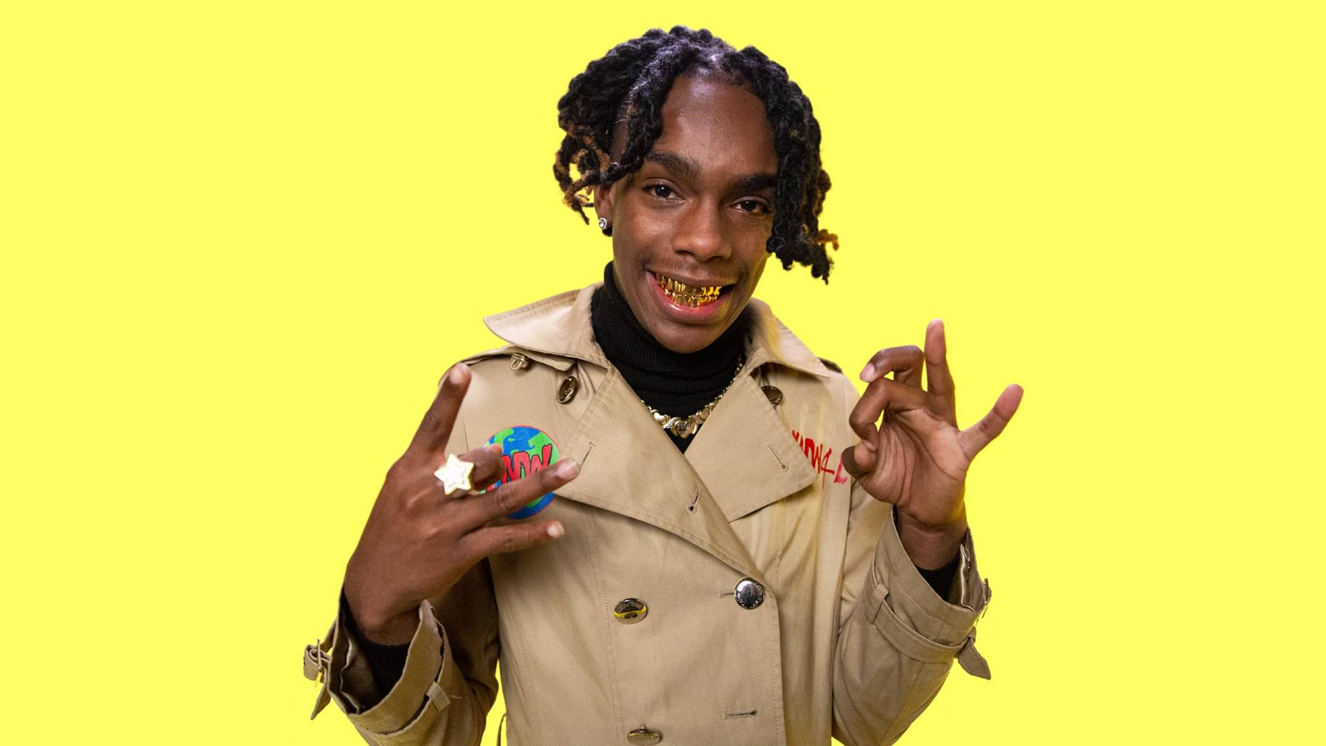 YNW Melly 出現在 Genius 節目中 壁紙 — 一位辮子頭髮和金色牙齒的年輕男子微笑著，手持地球儀並做出手勢，背景是明亮的黃色。