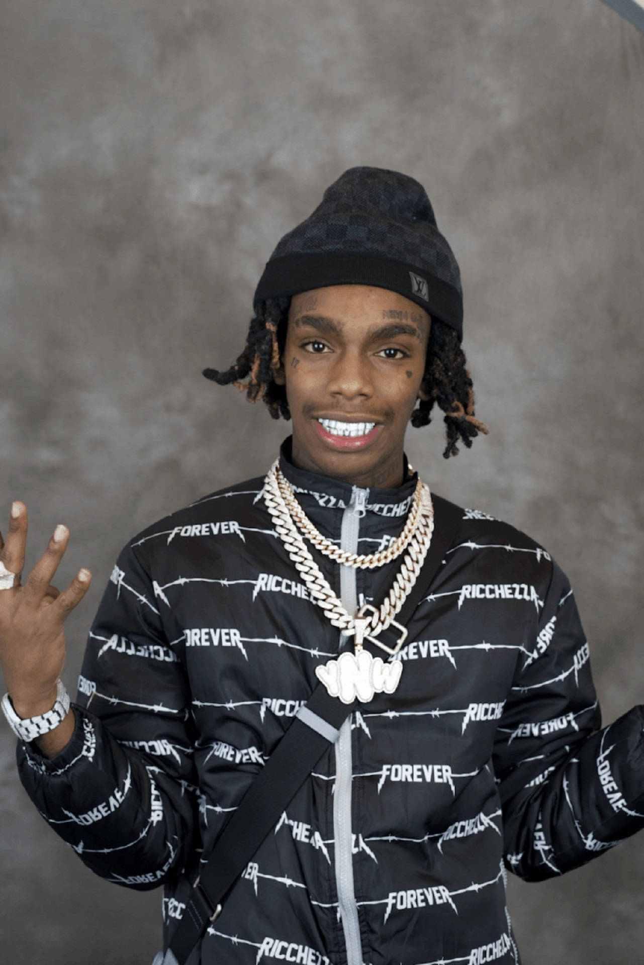 YNW Melly 嘻哈藝術家 壁紙 — 一位留著辮子頭、戴黑色冷帽的年輕男子微笑著，身穿帶有鐵絲網圖案和層疊金鏈的黑色外套。